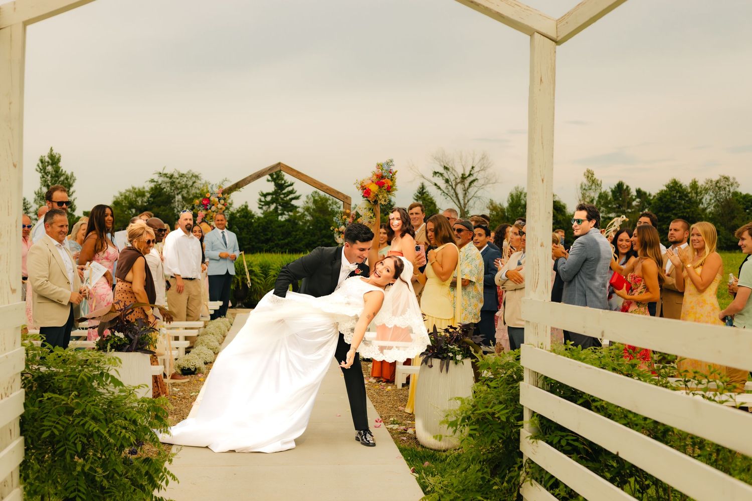 Groom dips bride for a kiss at outdoor wedding ceremony under white arbor with guests applauding.