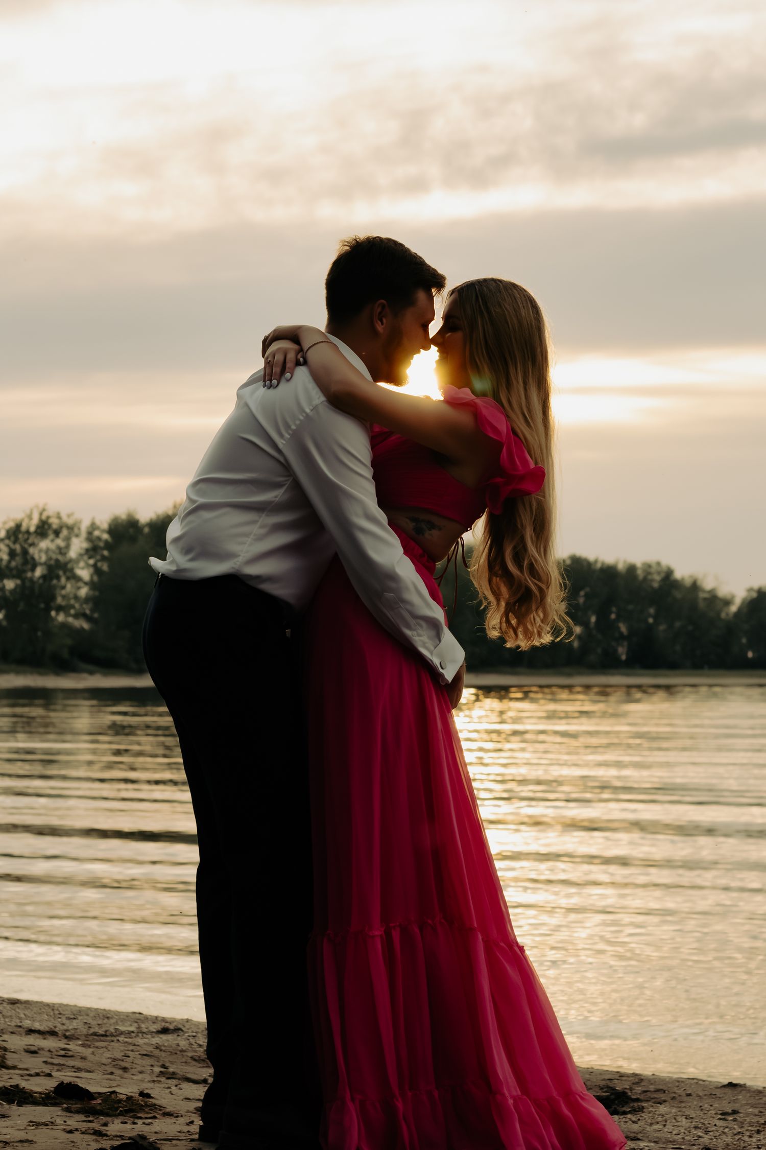 Romantic couple embracing and kissing on a sandy beach at sunset, woman in flowing pink dress, golden light reflecting on water.