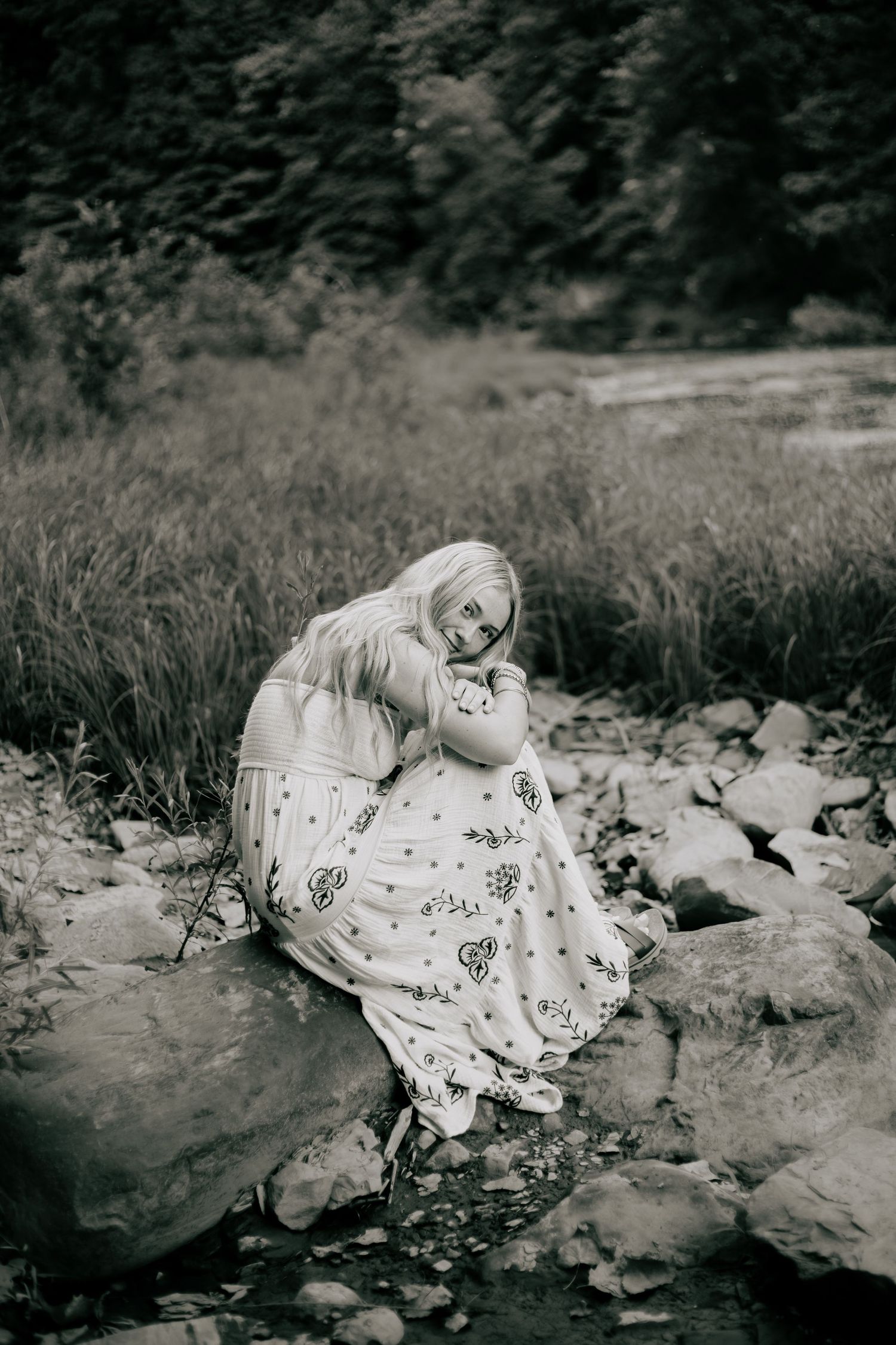 Black and white photo of a woman in a floral dress sitting on rocks by a stream in a lush forest.