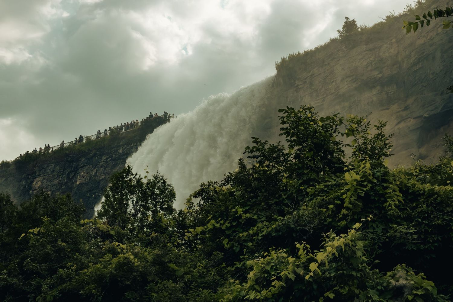 A massive waterfall cascades down a steep forested mountain slope under dramatic cloudy skies.