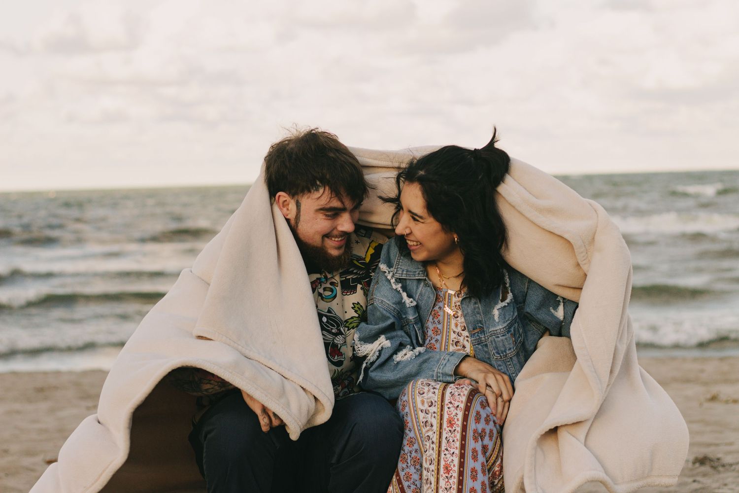 Romantic beachside photos showing tender moments wrapped in a cream colored blanket.