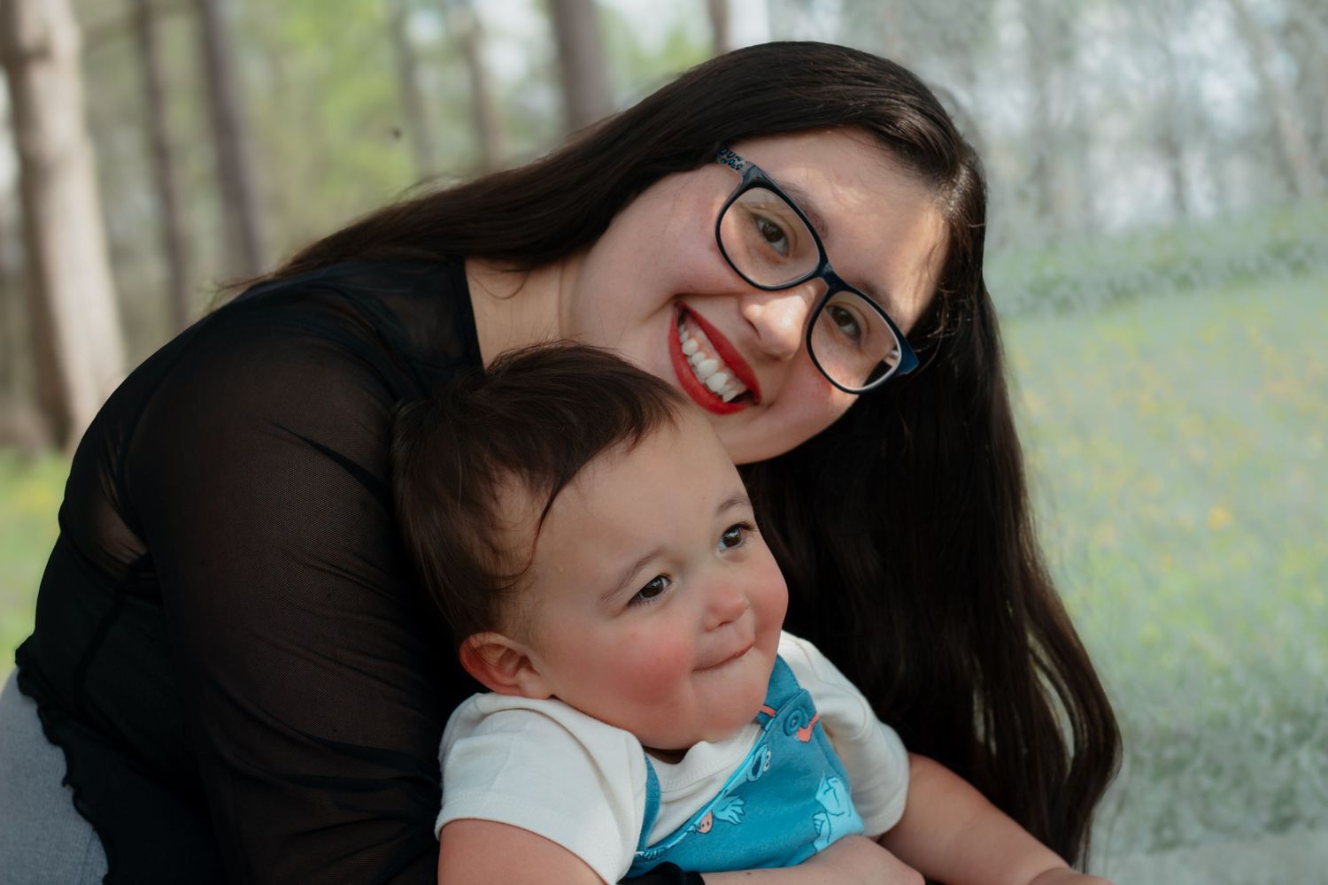 Young woman with glasses and red lipstick smiling while hugging an adorable baby outdoors.