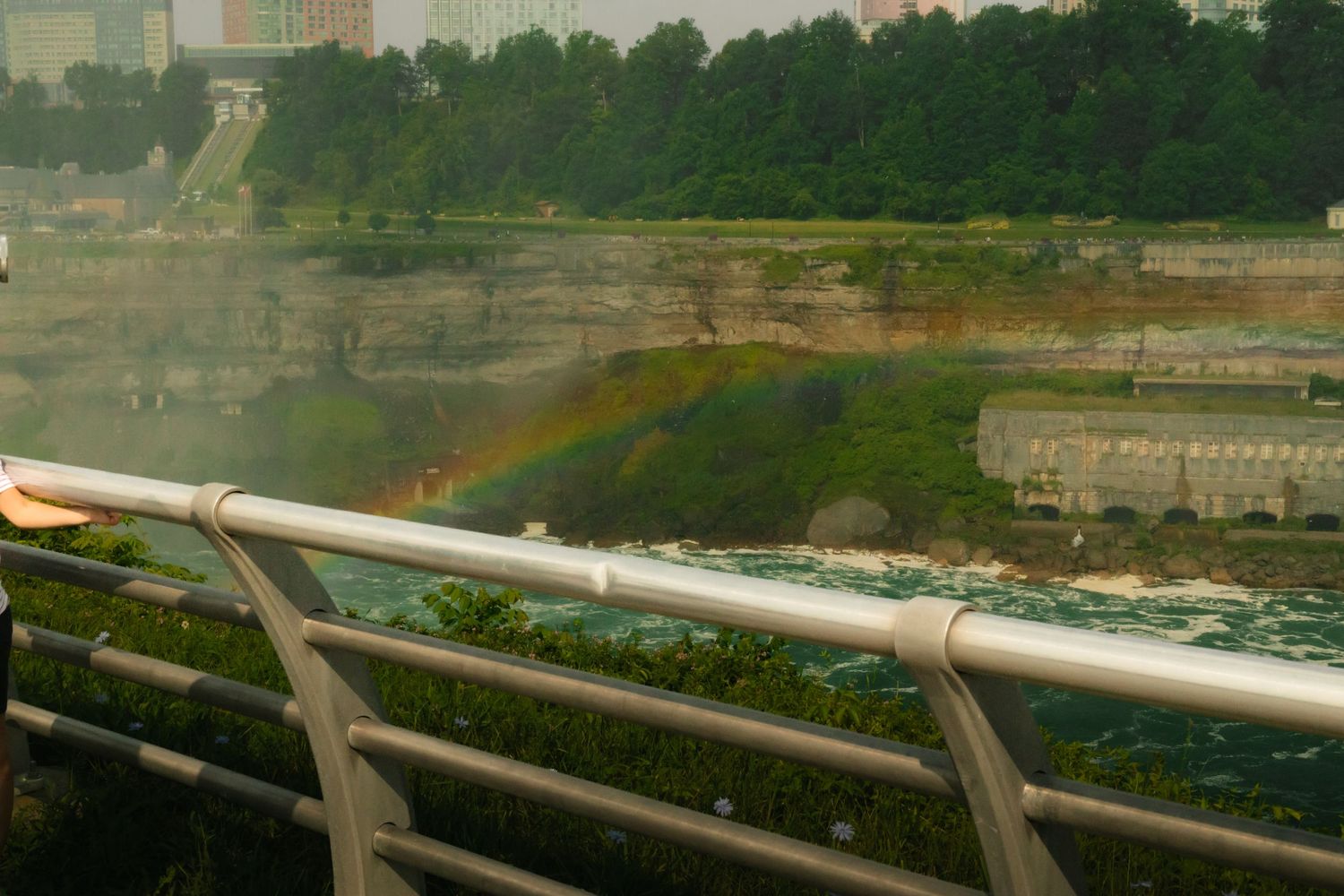 Rainbow over Niagara Falls viewed from a metal railing bridge with lush greenery and mist in the background.
