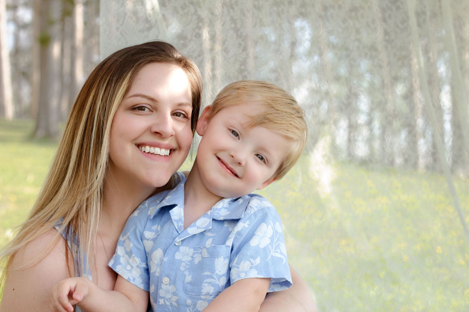 A smiling young woman hugs a happy blonde toddler boy in a blue shirt outdoors near trees.