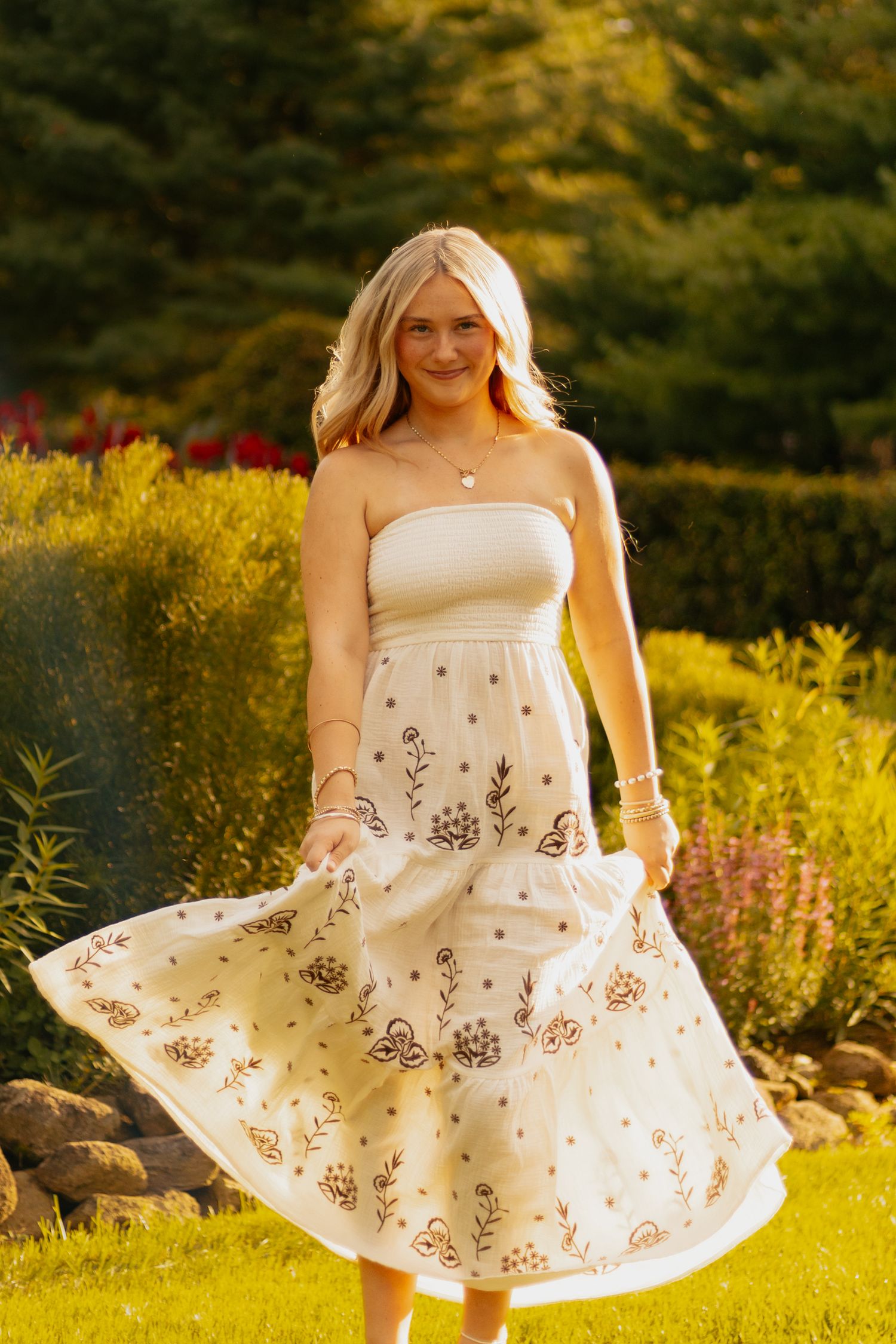 Young blonde woman twirling in a white floral embroidered midi dress outdoors in a lush garden at golden hour.