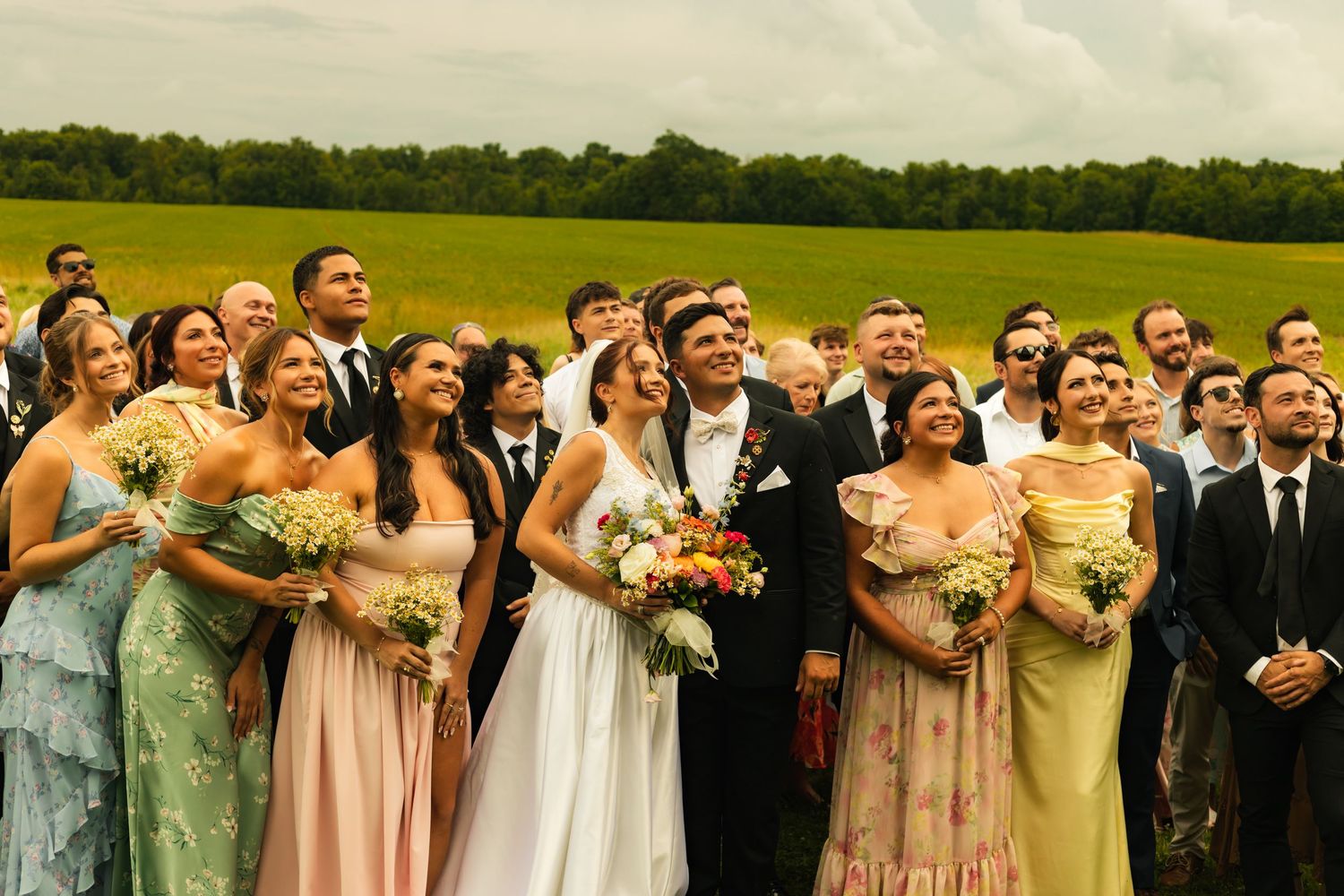 Wedding party smiling outdoors in a green field, bride in white gown surrounded by colorful bridesmaids and groomsmen.