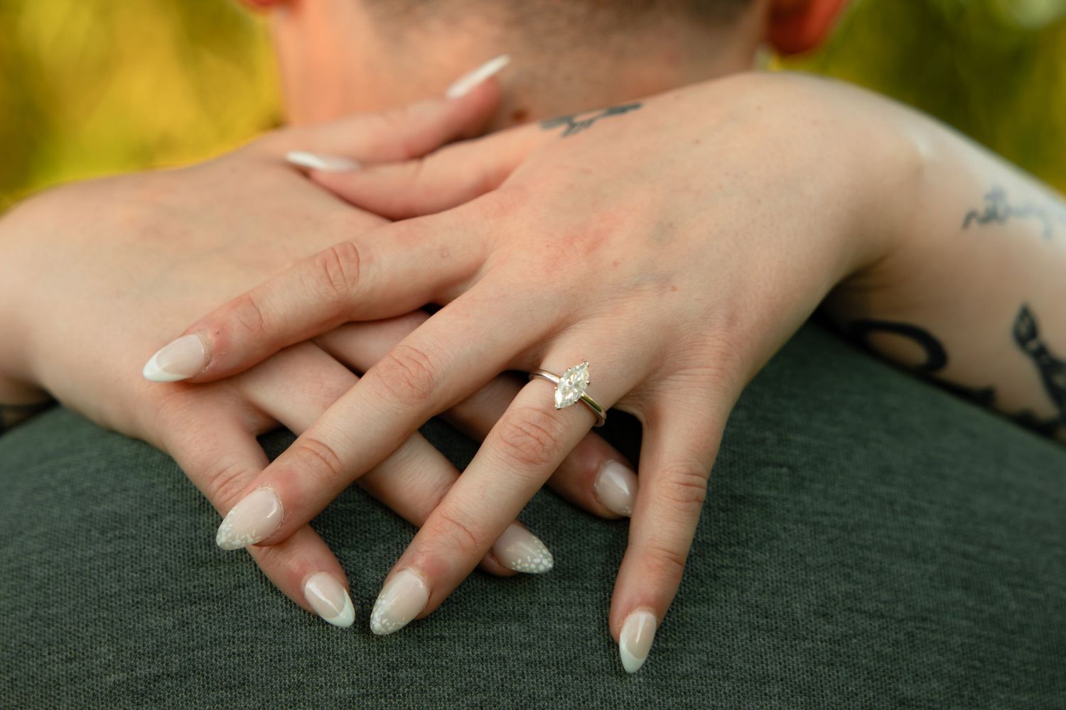 Close-up of a woman's hand with white nails and a diamond engagement ring resting on a man's shoulder.