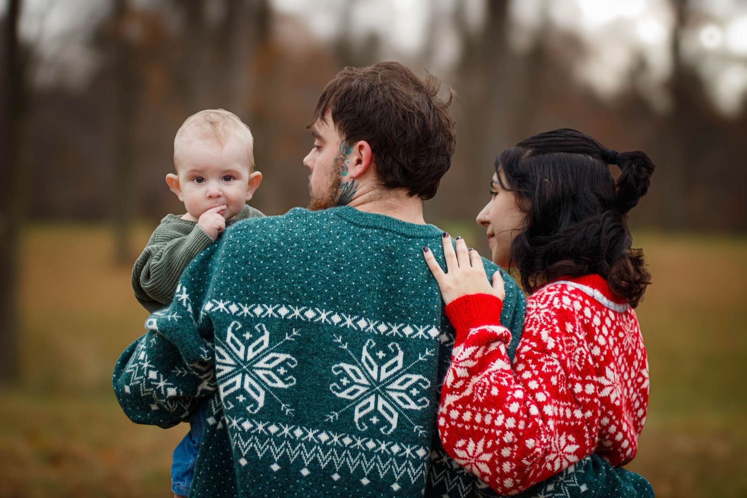 Family wearing festive holiday sweaters looks lovingly at their baby outdoors in autumn.