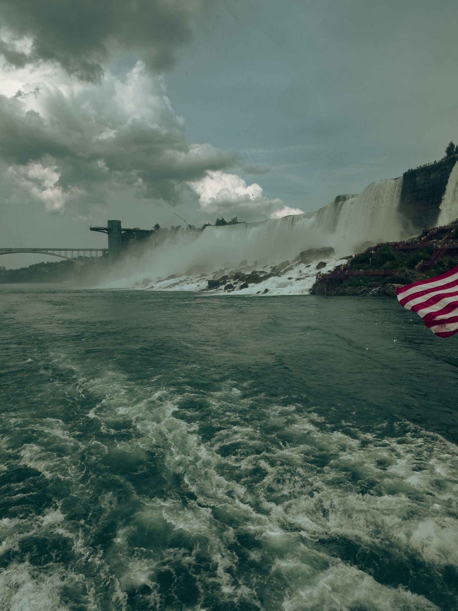 American flag waves from a boat near Niagara Falls with churning waters and dramatic cloudy skies in the background.