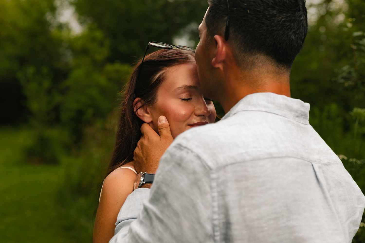 A couple shares a romantic moment outdoors, with the man gently touching the woman's face near lush green trees.