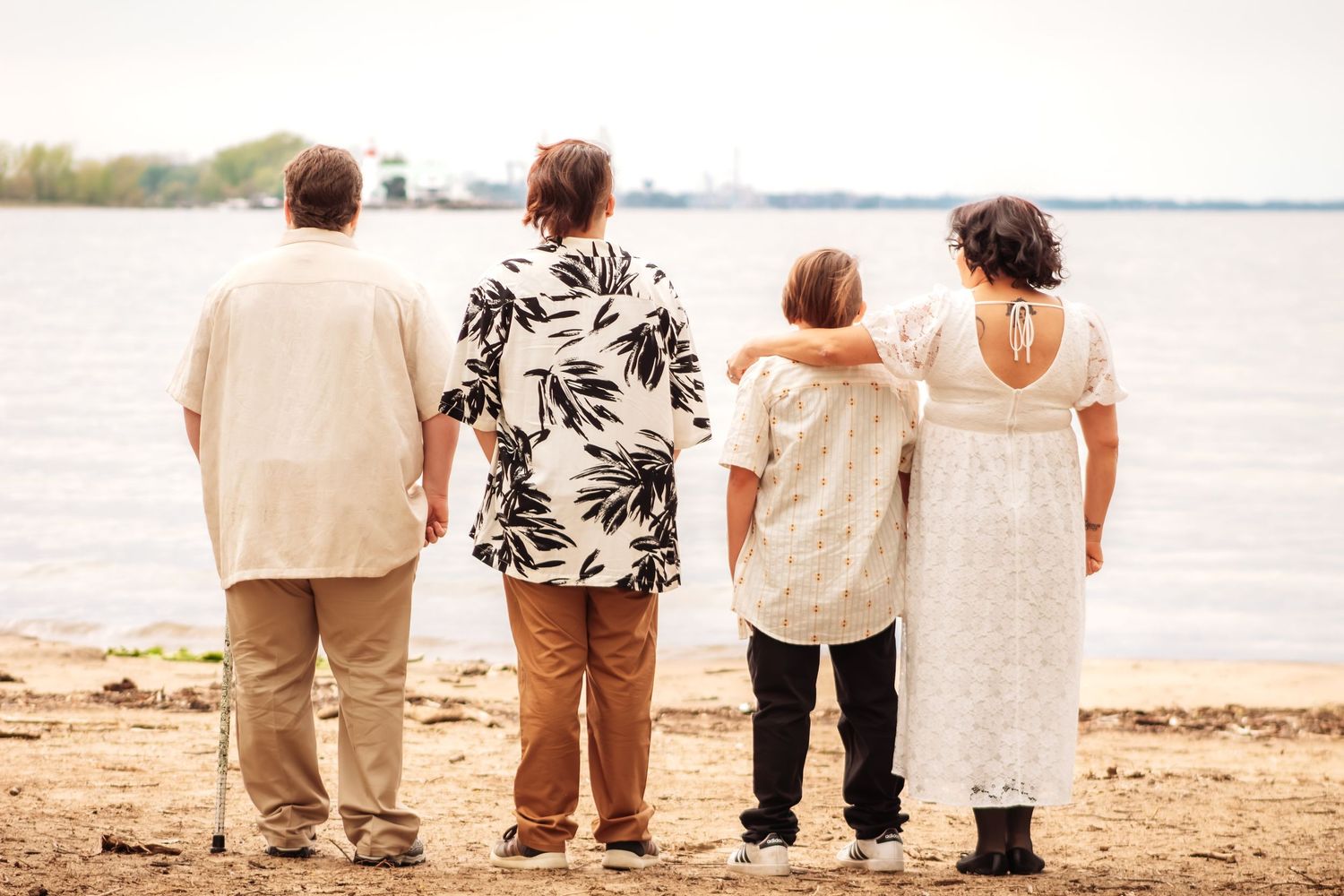 Four people standing together on a sandy beach shore, viewed from behind, looking out over calm water.