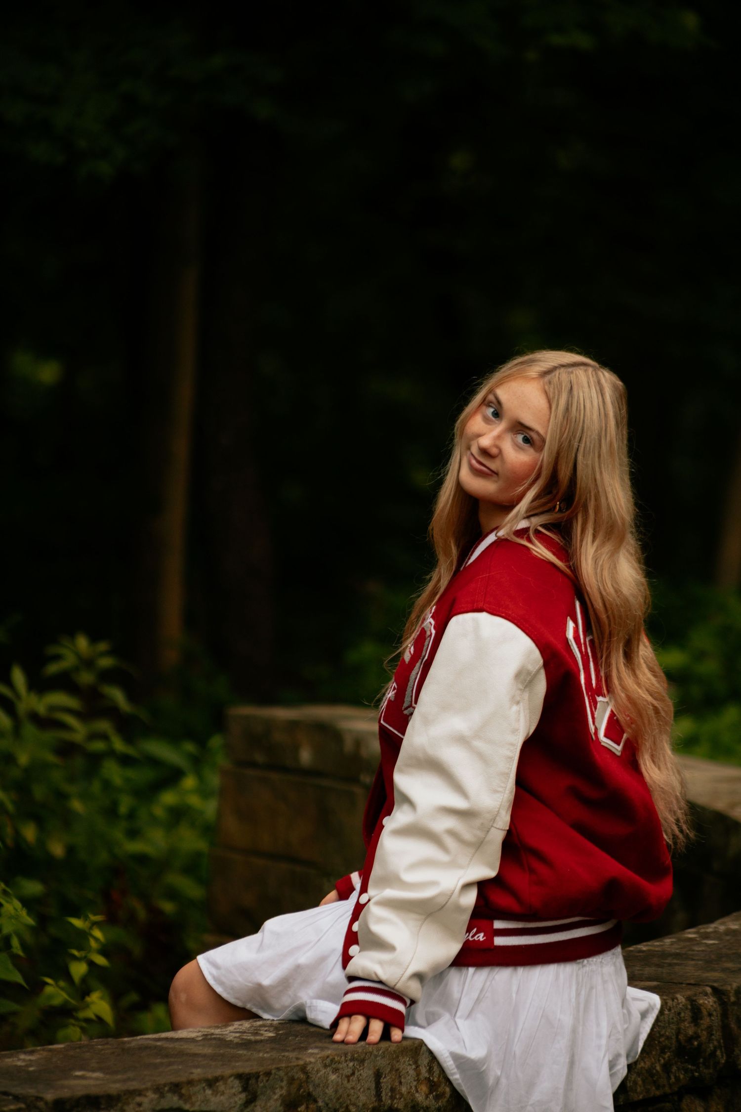 Young blonde woman in red varsity jacket and white skirt sitting on stone wall in lush green forest setting.