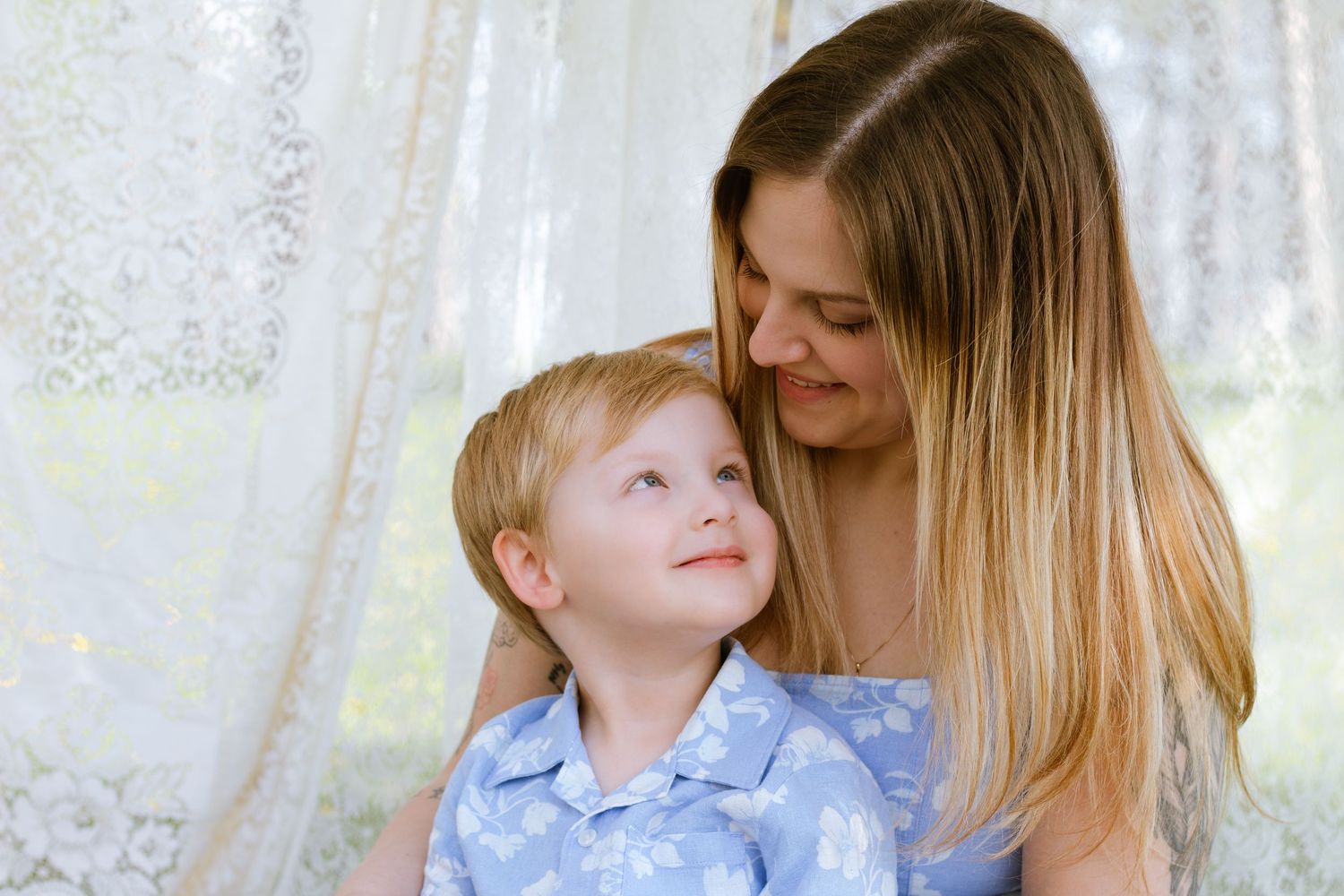 A loving mother with long blonde hair smiles at her young blond son in a blue floral shirt near lace curtains.