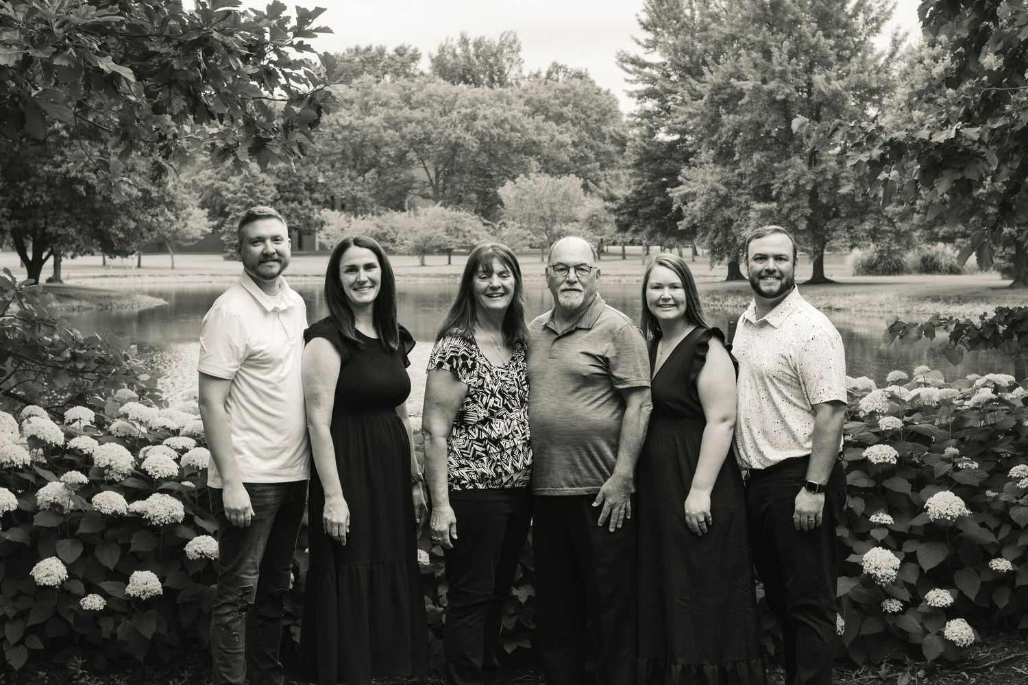 Black and white family portrait of seven smiling adults posing outdoors near a pond surrounded by hydrangeas and trees.