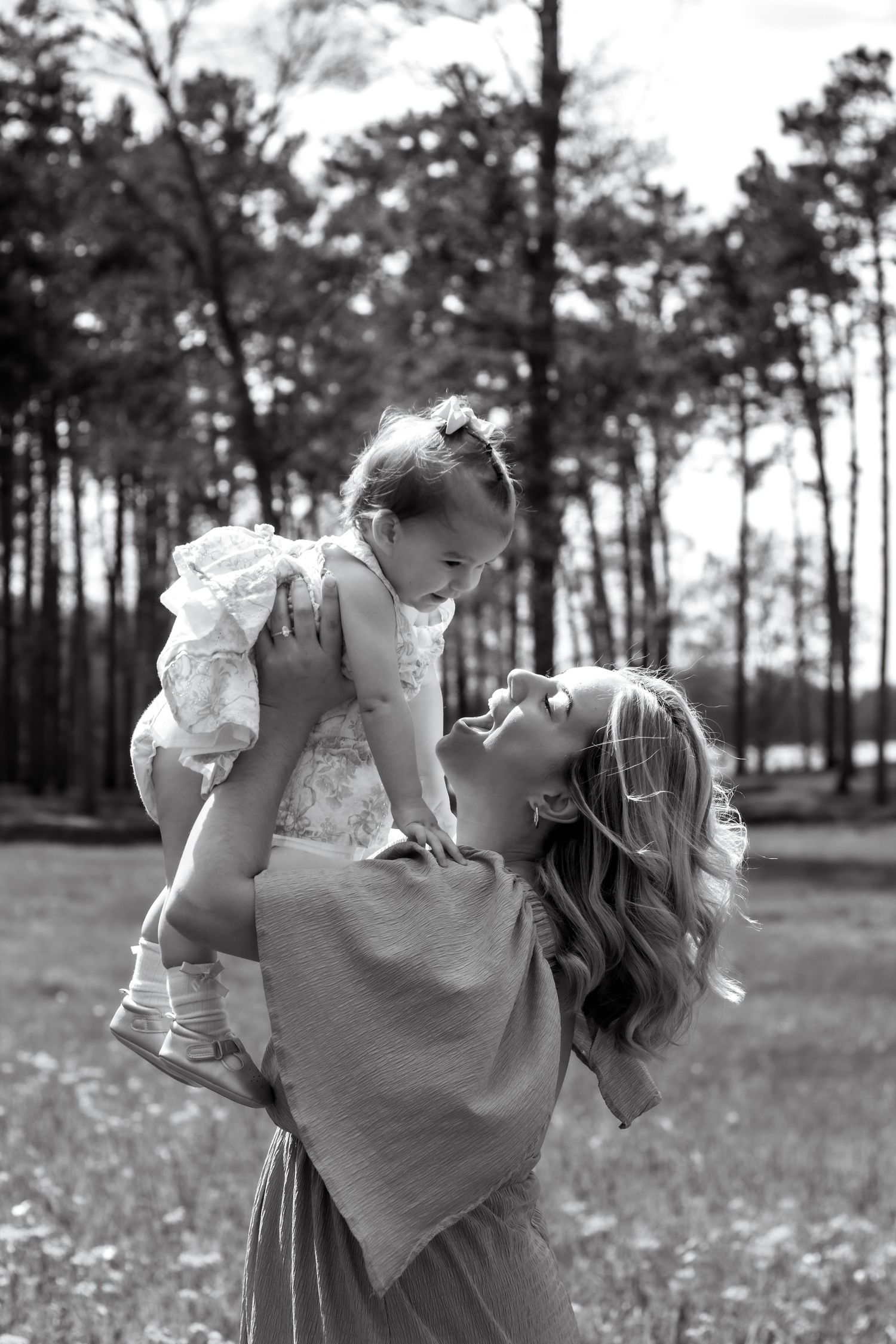 Black and white photo of a smiling mother lifting her baby girl in a sunny outdoor meadow surrounded by tall trees.
