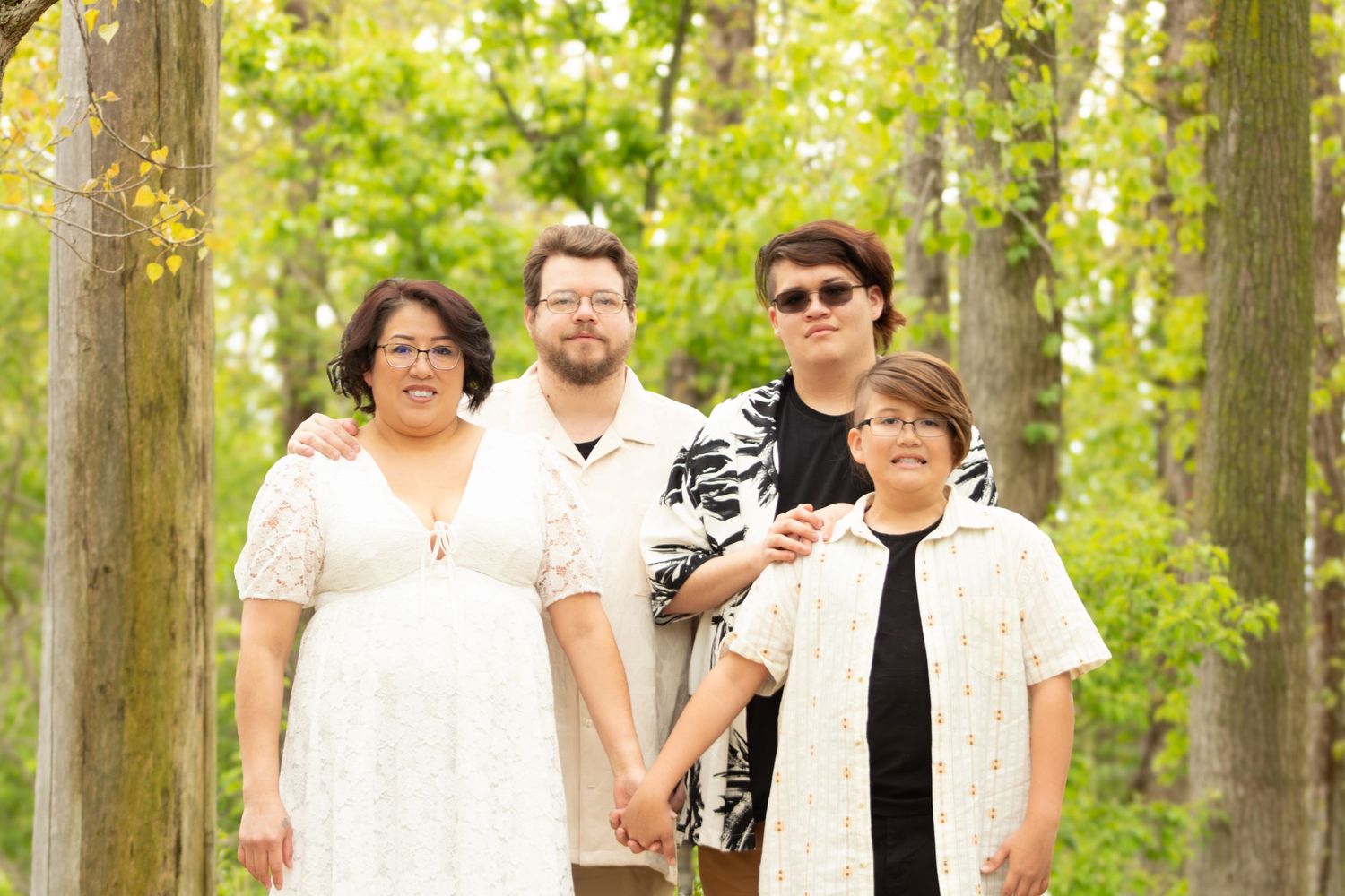 Family of four posing outdoors among tall trees, wearing white and black outfits for a formal portrait.