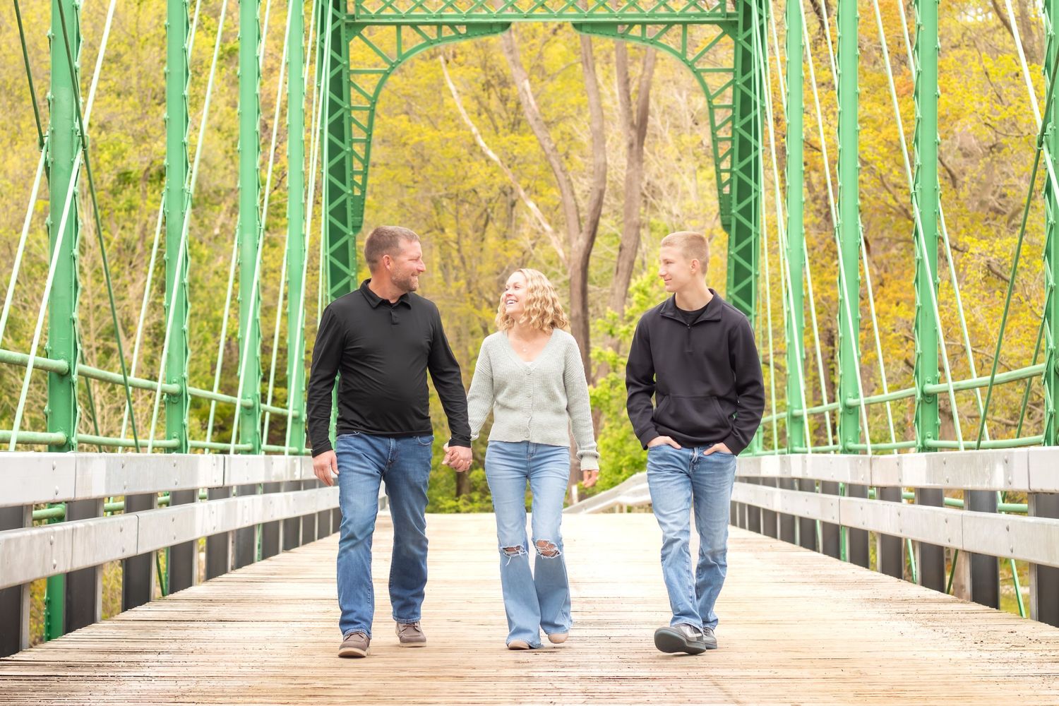 Family of three walking on a green iron bridge surrounded by autumn trees, smiling and holding hands.
