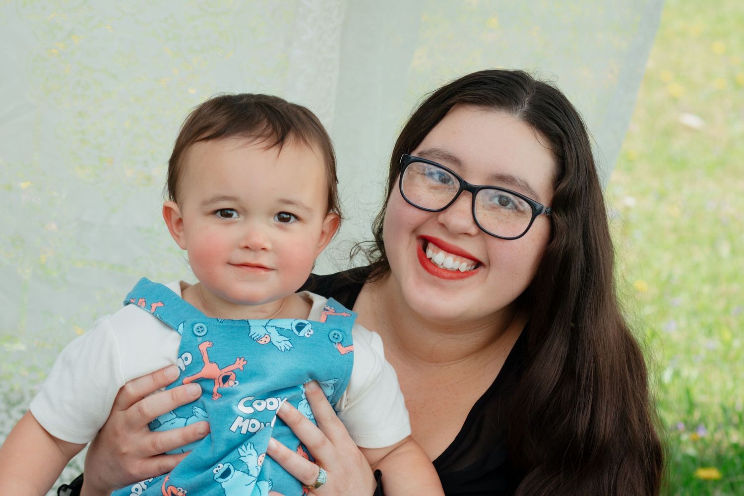 A smiling woman with glasses holds a happy baby in a blue outfit outdoors.