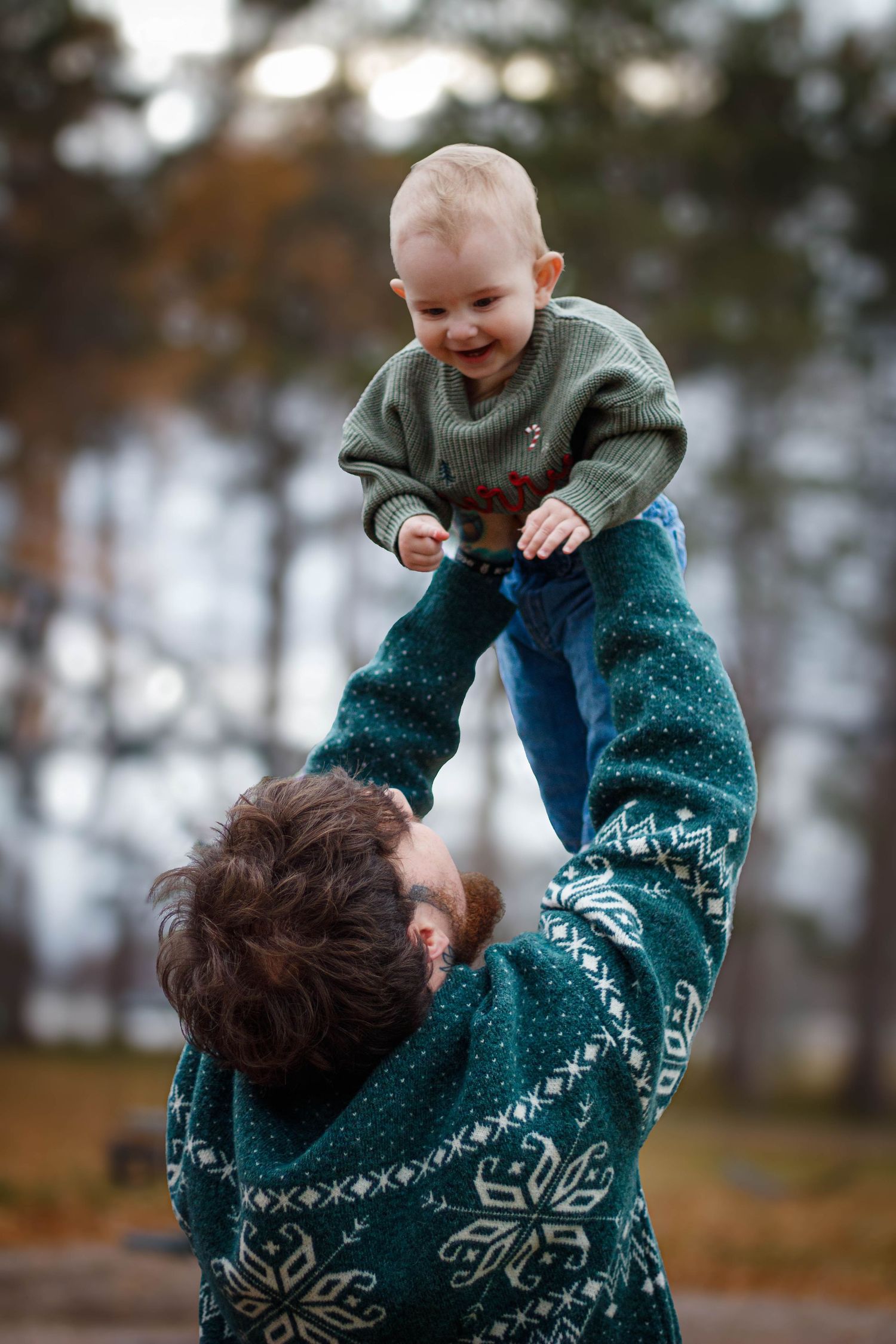 Parent lifts laughing baby high in the air while wearing matching festive winter sweaters.
