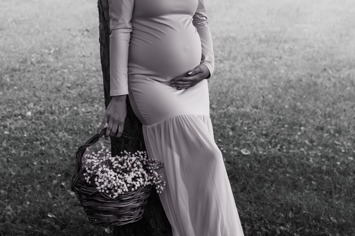 Black and white photo of a pregnant woman in a flowing dress holding a basket of wildflowers outdoors.