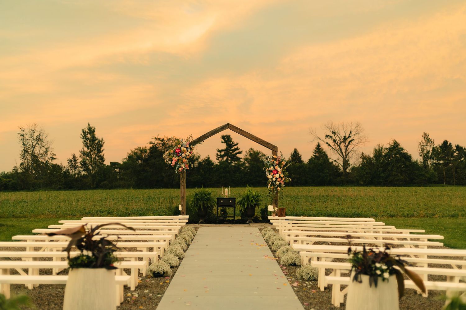Outdoor wedding ceremony setup with white benches, floral arch, and a aisle at golden hour sunset.