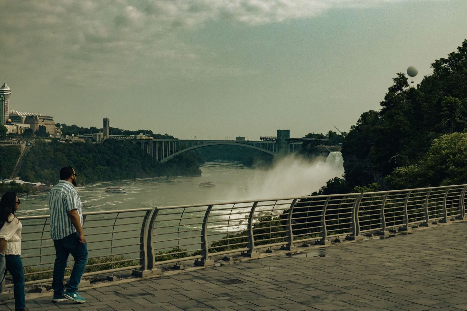 Two people stand on a promenade overlooking Niagara Falls with a bridge and mist in the background.