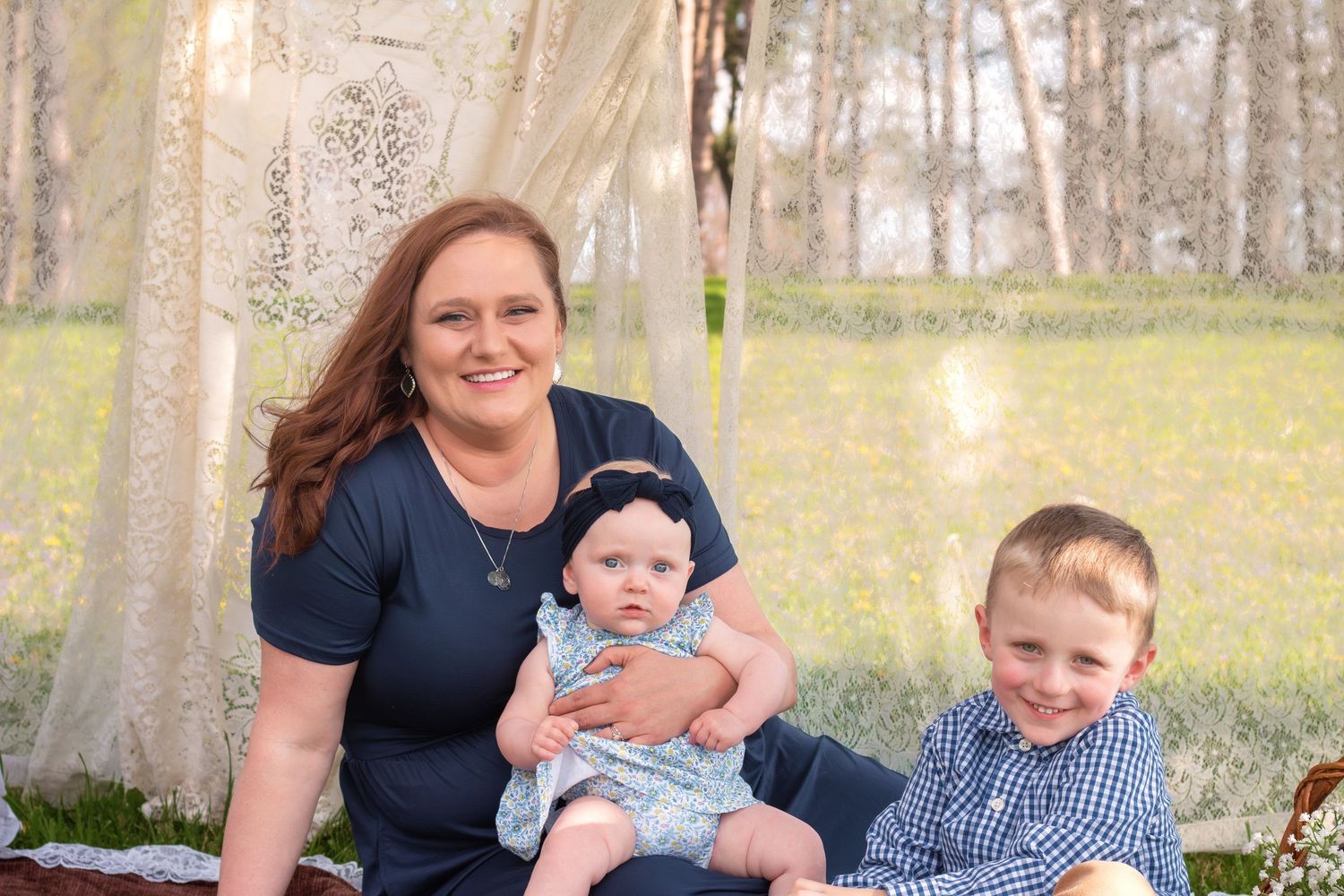 A smiling woman poses outdoors with her baby girl and young son in a charming rustic garden setting.