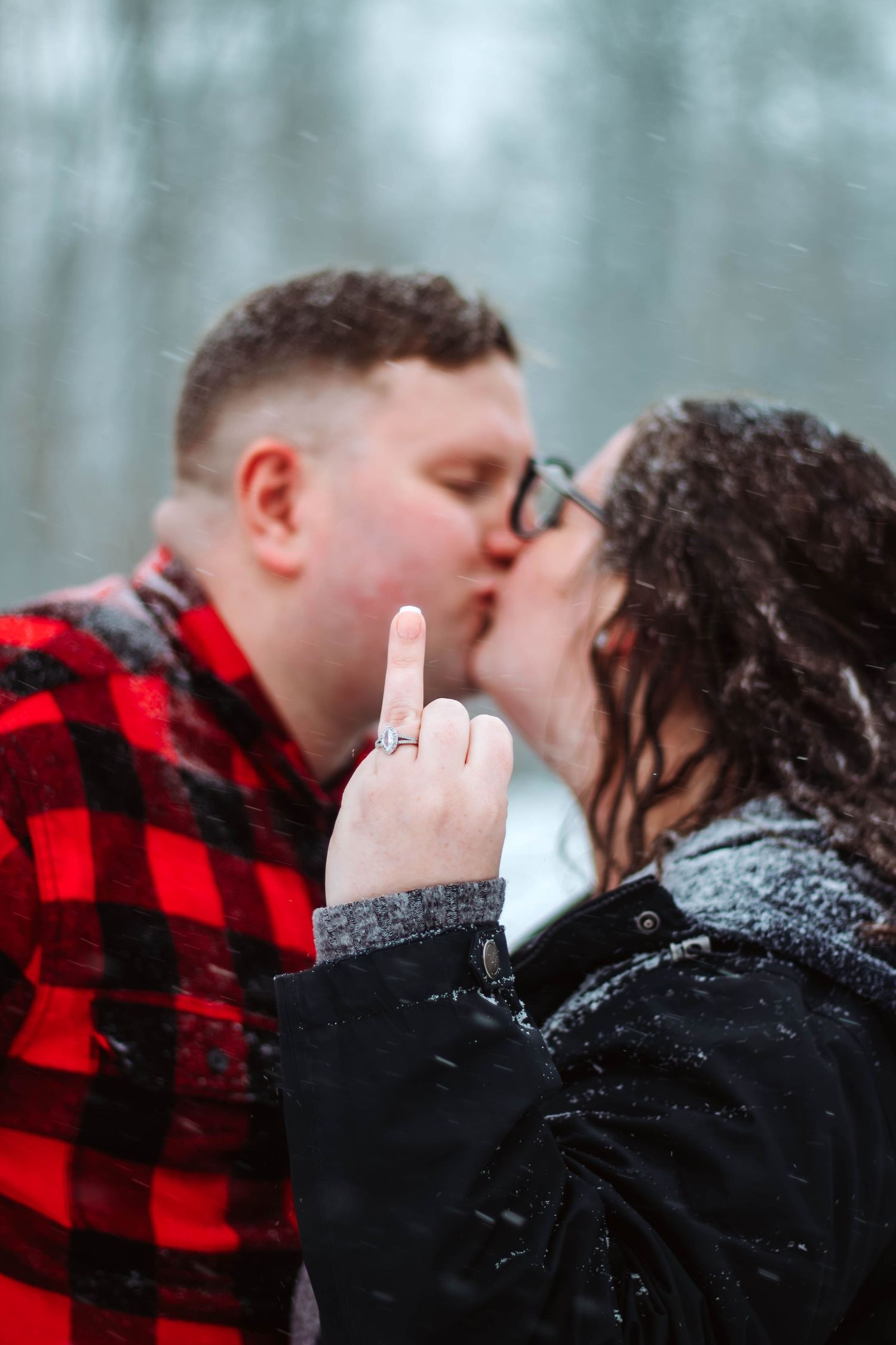 A couple sharing a romantic kiss in the snow while showing off an engagement ring.