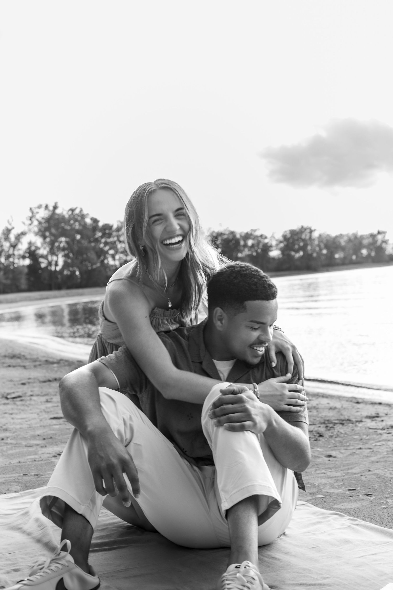 Black and white photo of a laughing couple sitting together on a beach blanket by a lake, sharing a joyful moment.
