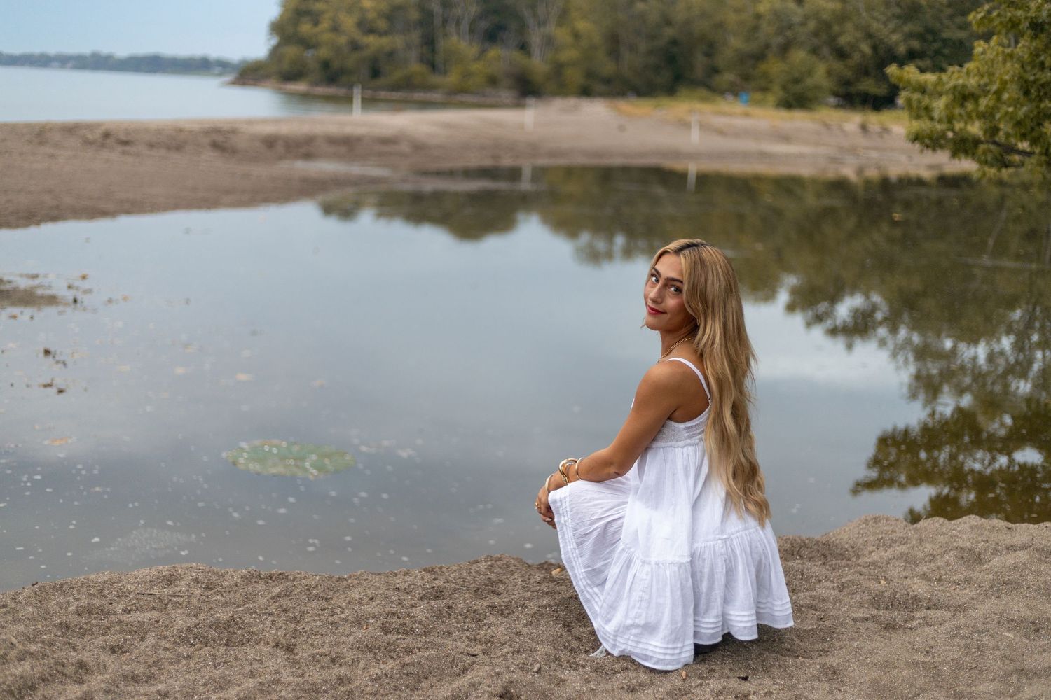 A blonde woman in a white dress sits gracefully on a sandy riverbank, gazing at the calm reflective water.