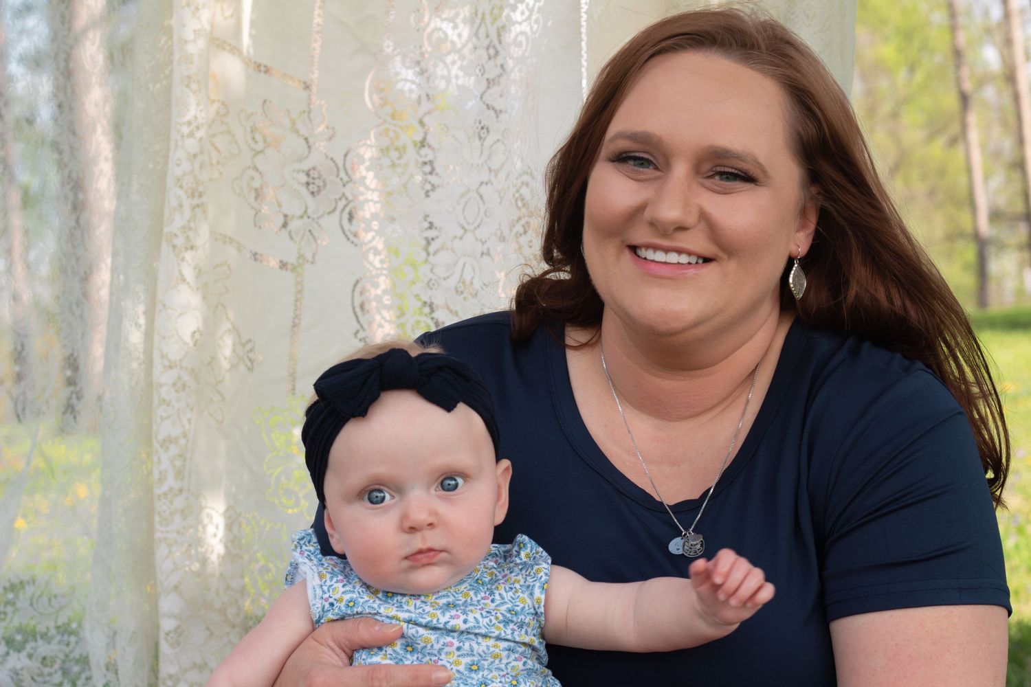 A happy mother smiles holding her infant baby girl wearing a black headband during an outdoor photo session.
