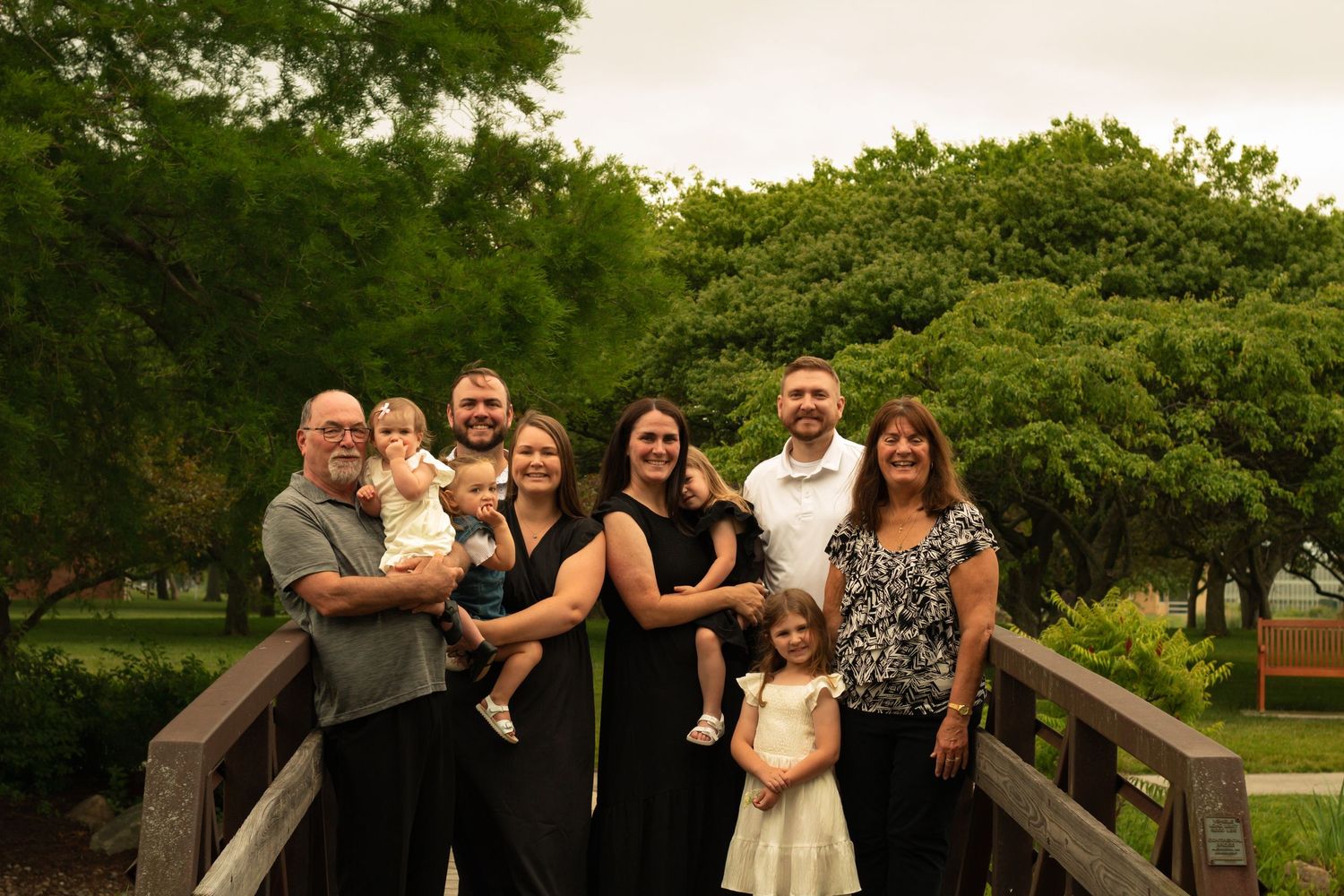 Family portrait of eight people in black attire posing on a wooden bridge surrounded by lush green trees.