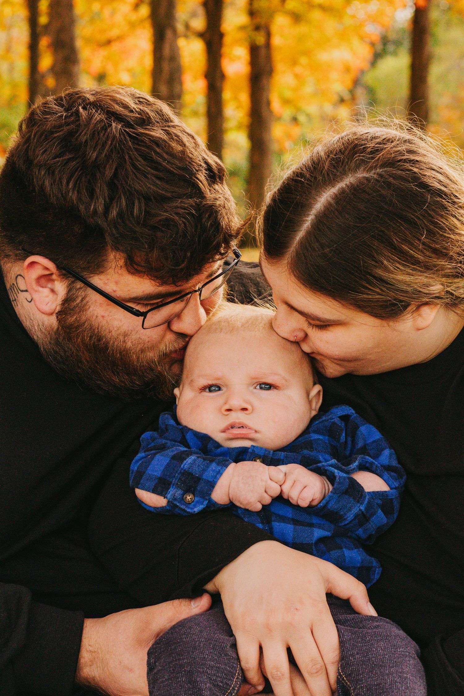 A tender family moment in an autumn forest as parents kiss their baby wearing a blue plaid shirt.