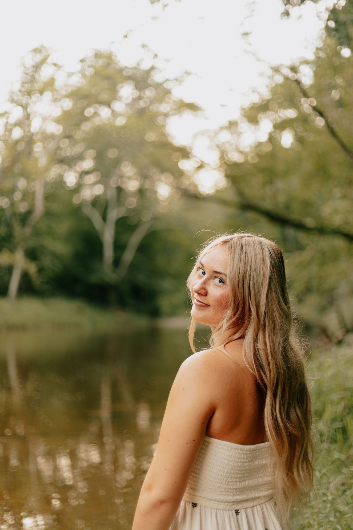 Young blonde woman in strapless dress smiling over shoulder near a sunlit forest pond.
