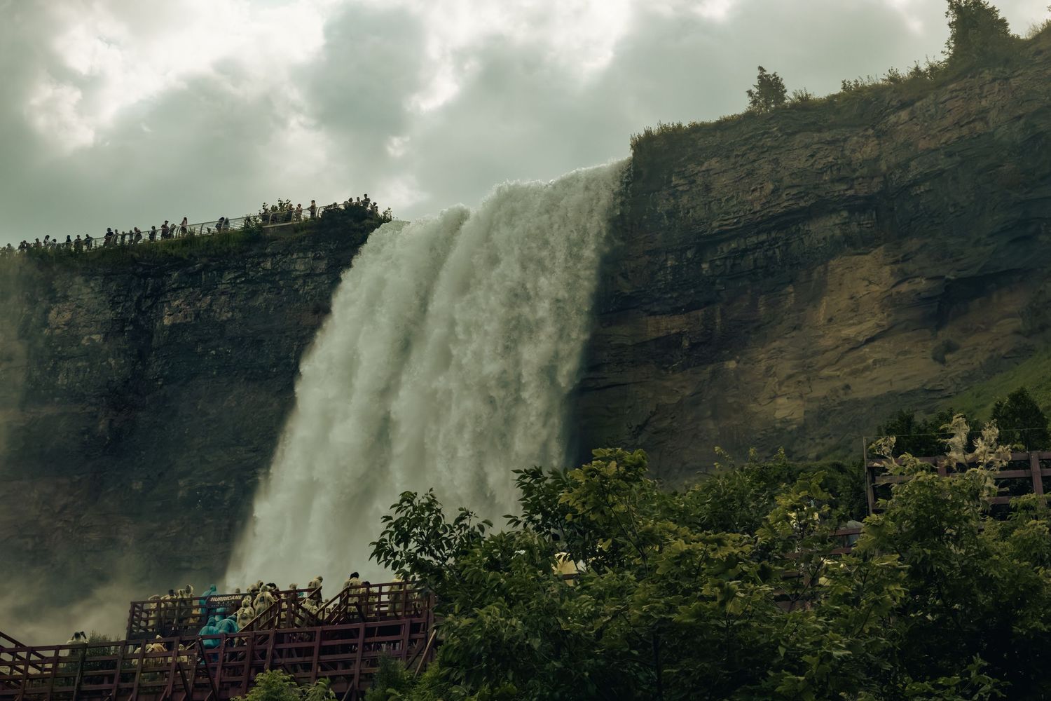Massive waterfall cascading down rocky cliffs with lush green trees and viewing platform below under cloudy skies.
