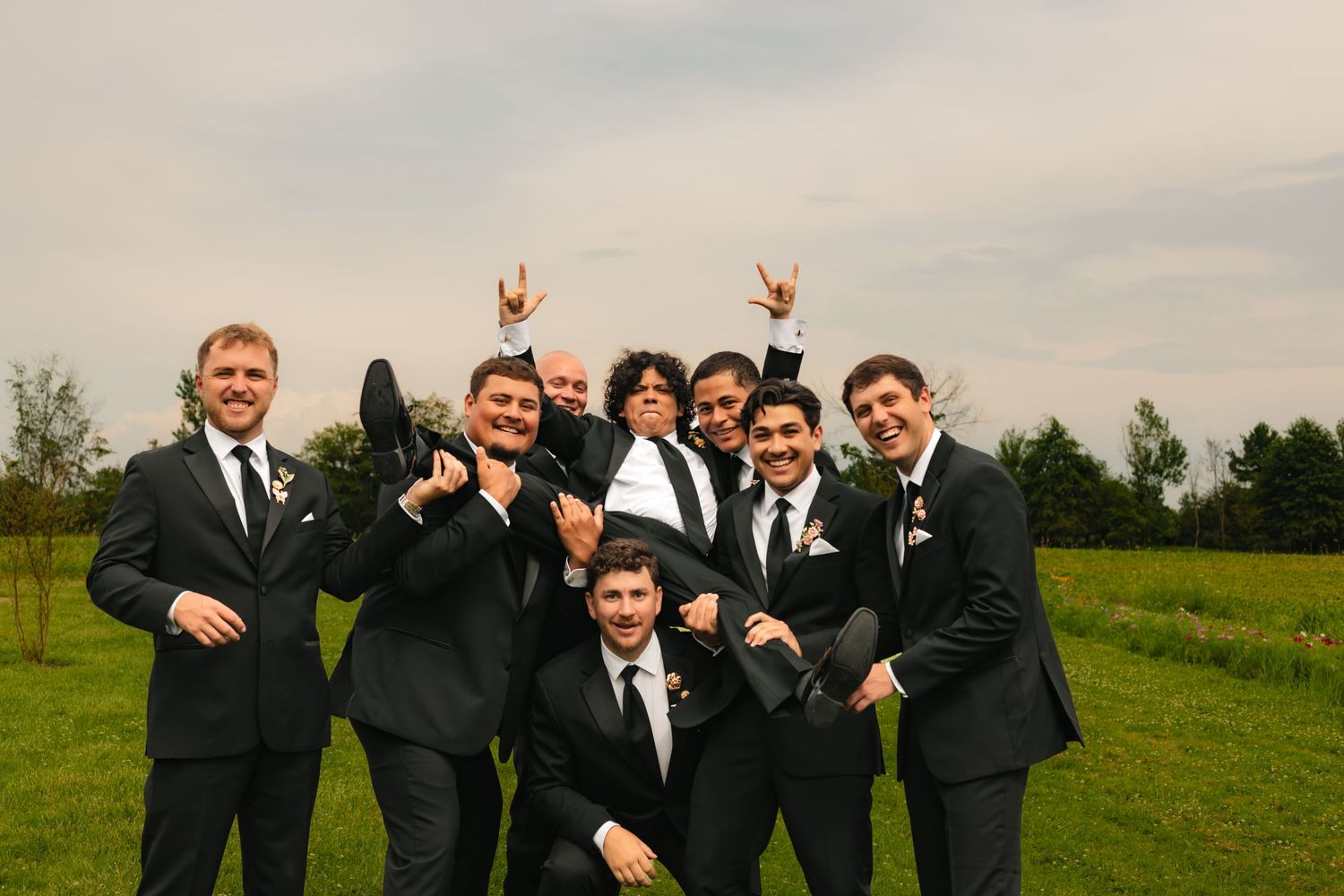 Group of groomsmen in black suits posing playfully on green lawn at outdoor wedding celebration.