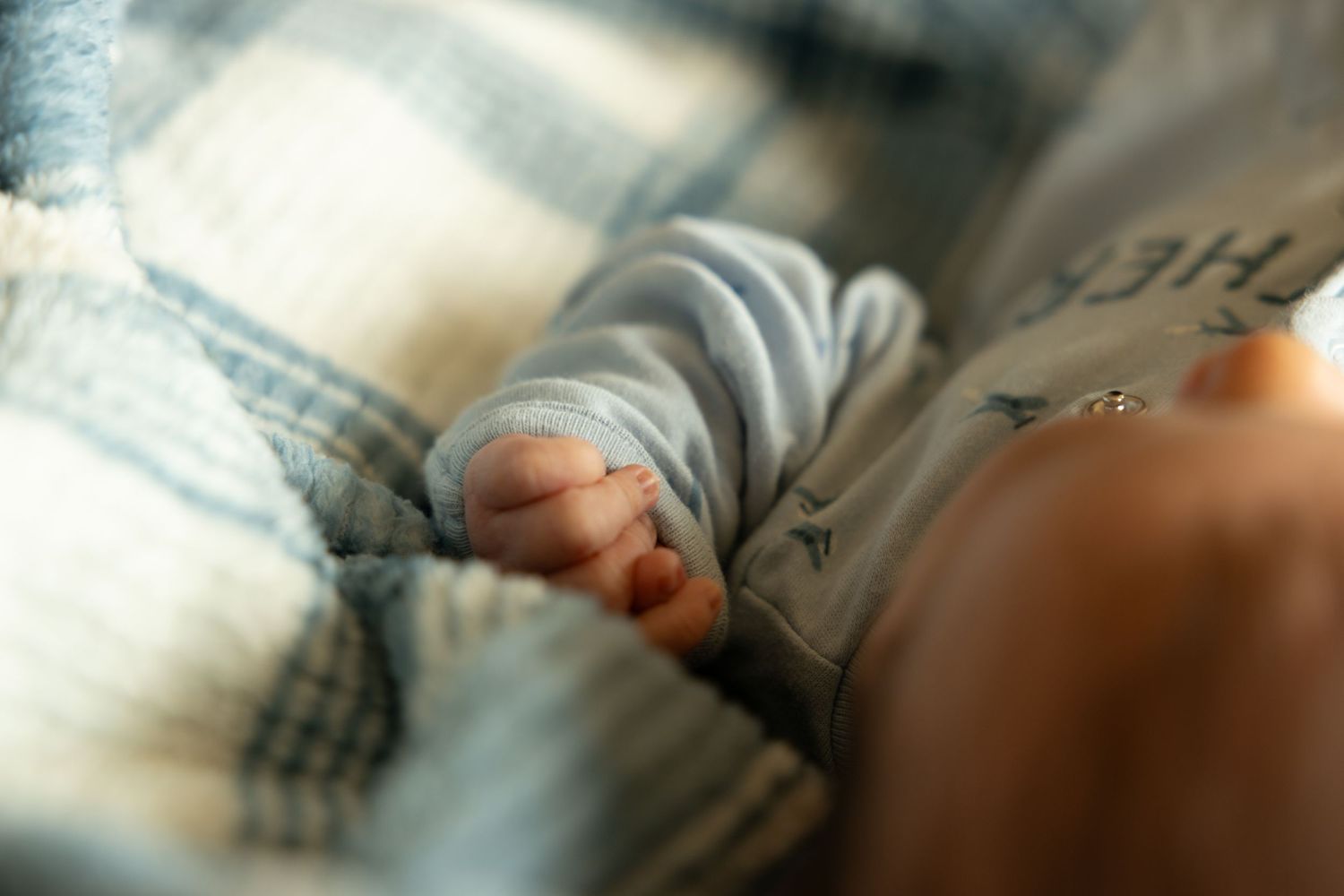 Close-up of a newborn baby's tiny hand in a soft blue onesie, resting peacefully on a plaid blanket.