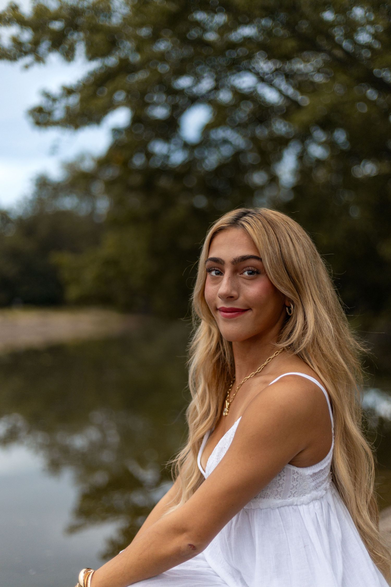 Young blonde woman in white dress smiling by a serene lake with lush green trees in the background.