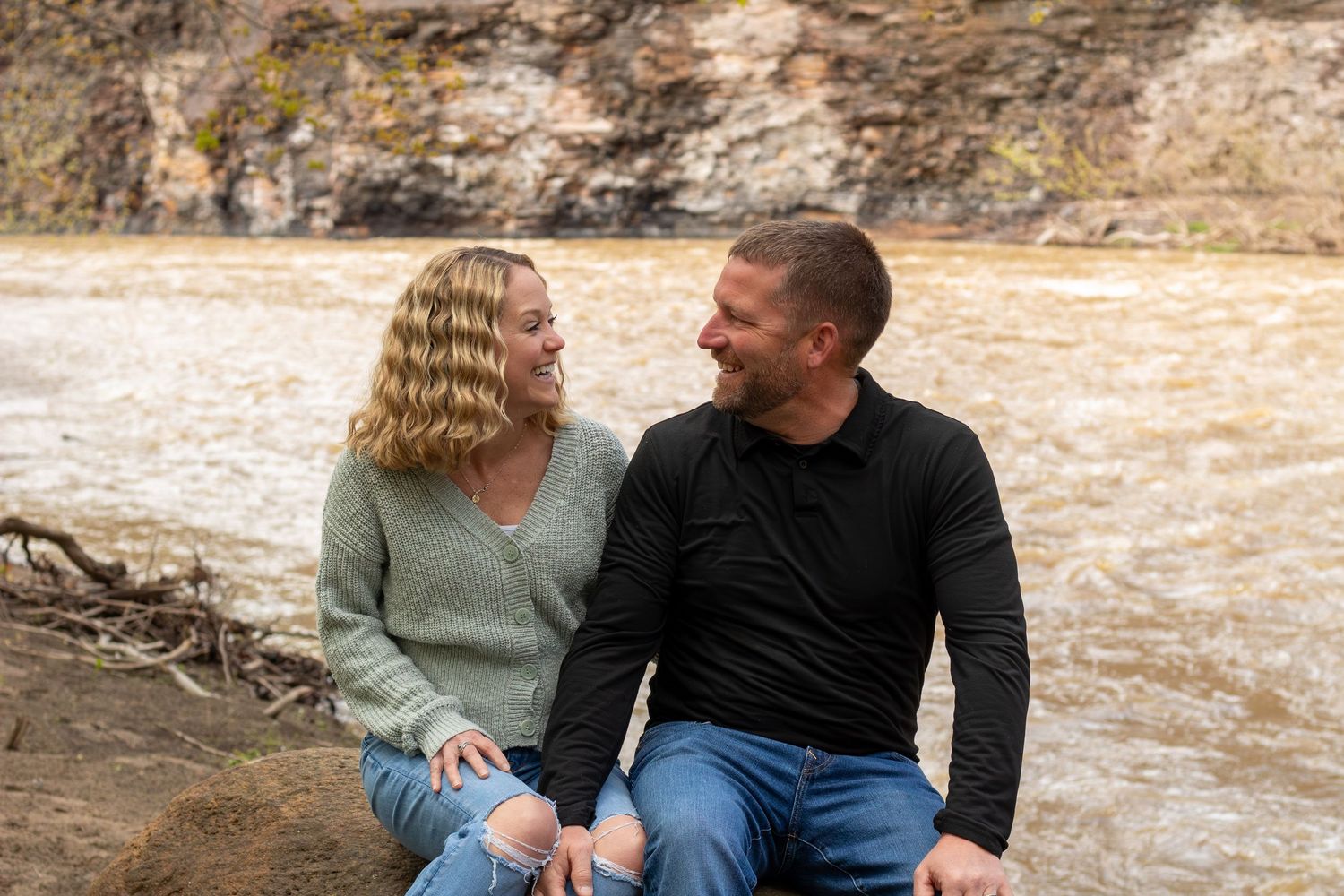 Couple sitting together smiling by a rushing river with rocky cliffs in the background outdoors.