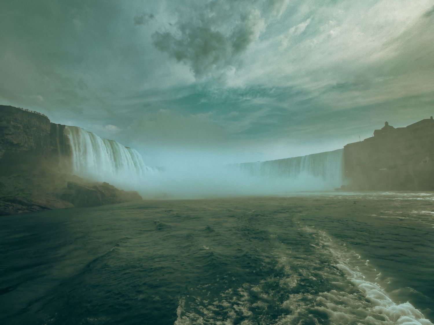 Dramatic waterfall cascading into misty turquoise waters under a stormy sky with towering cliffs on both sides.