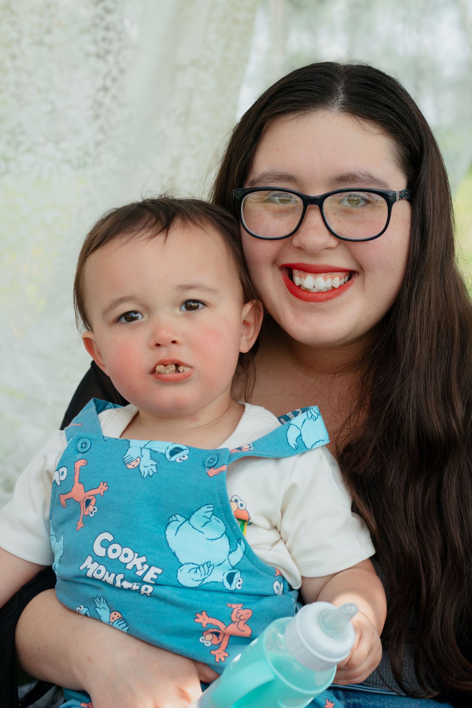 Young woman with glasses and red lipstick smiling while holding a baby wearing a Cookie Monster bib.