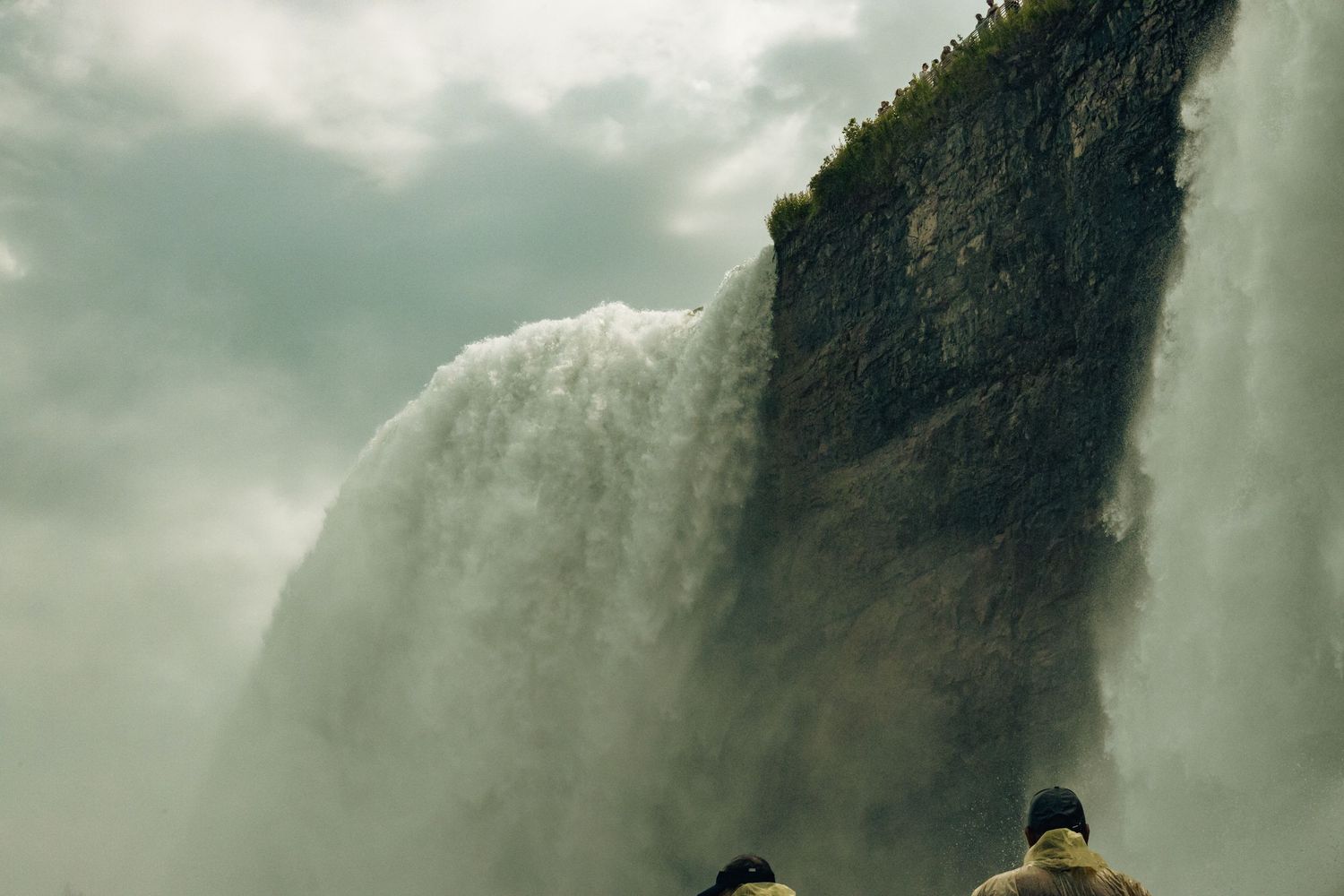 Two people stand in awe before a massive waterfall cascading down a dramatic cliff face into misty clouds below.