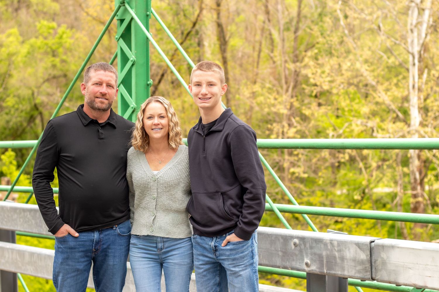 Family of three smiling on a green bridge surrounded by lush trees in a warm outdoor portrait.