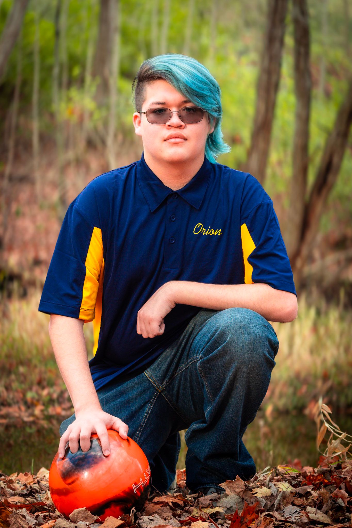Person in blue uniform crouching near fallen leaves while holding a bright orange pumpkin.