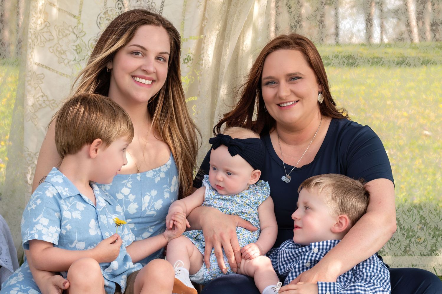 Two women pose with three young children including a baby girl during an outdoor family portrait session.