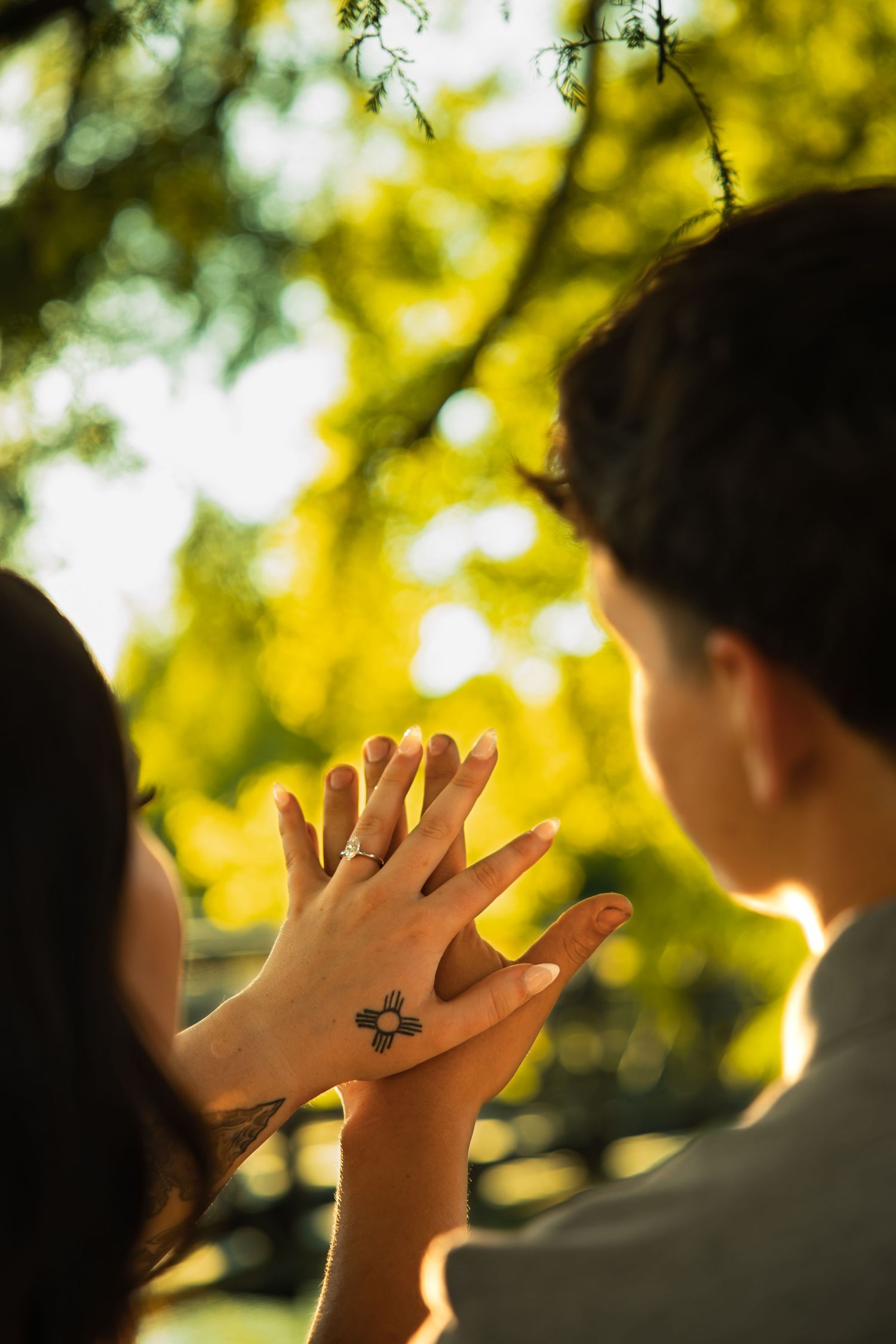 Couple holding hands outdoors with a New Mexico Zia sun tattoo visible, surrounded by lush green trees in warm sunlight.