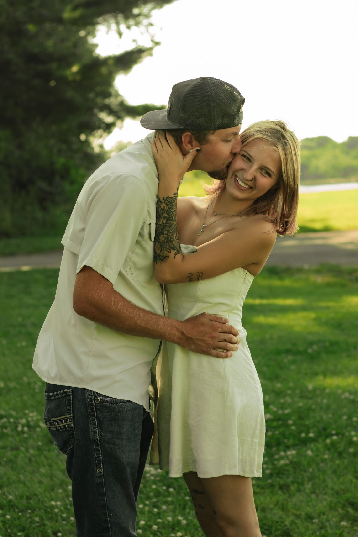 Young couple sharing a romantic kiss in a green park, man in snapback cap with tattoo embracing smiling blonde woman in white dress.
