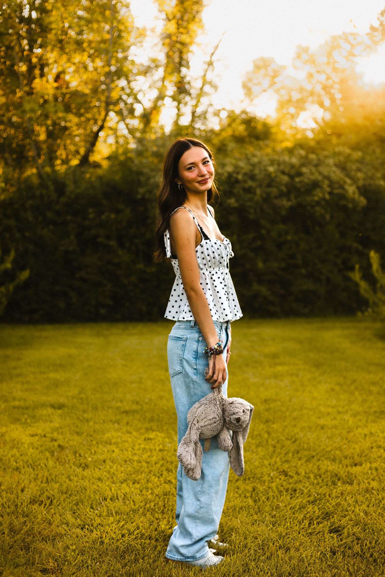 Young woman in polka dot top and jeans holds a stuffed bunny while standing on green grass at golden hour.