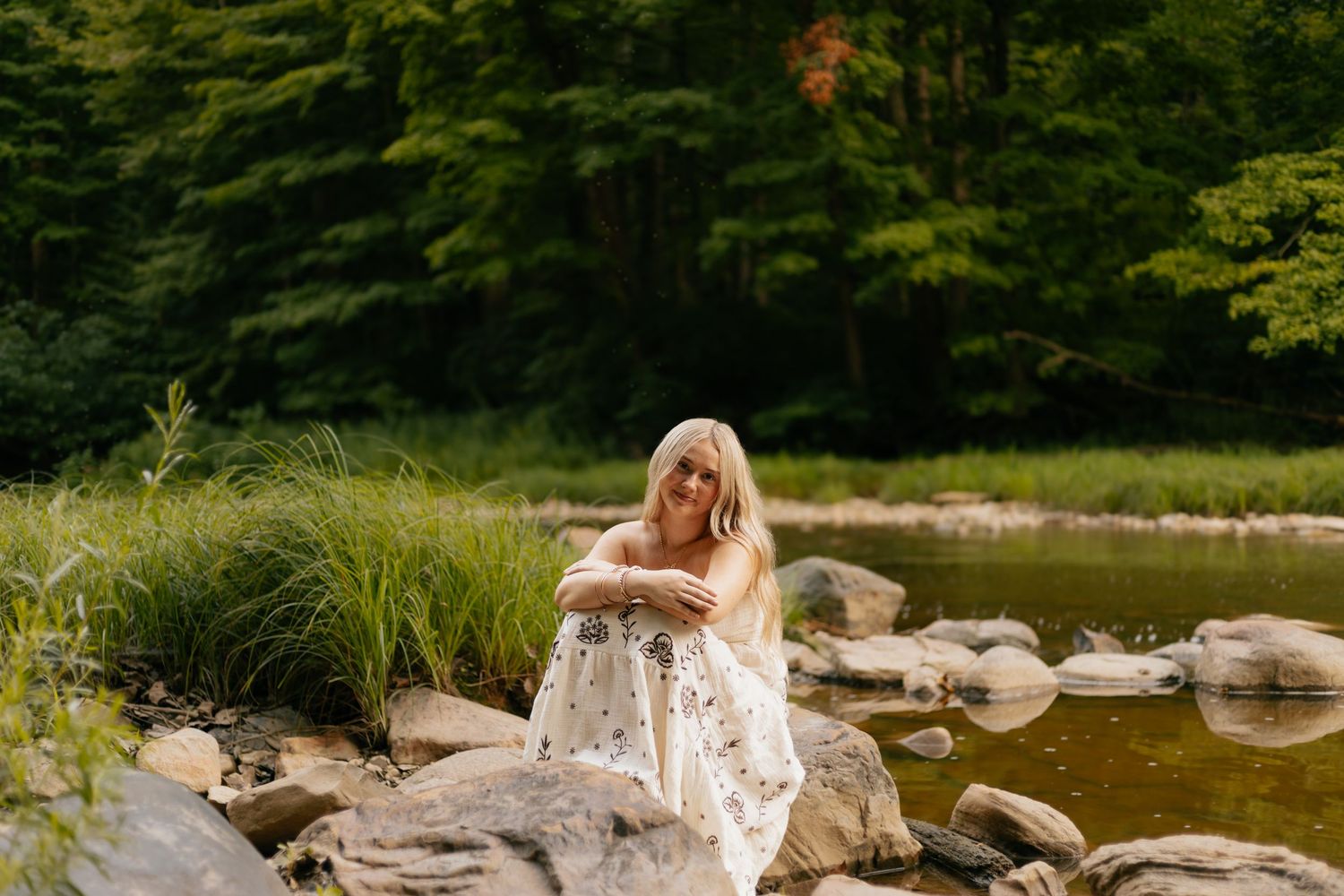 Woman in floral dress sitting on rocks by a serene river surrounded by lush green forest.
