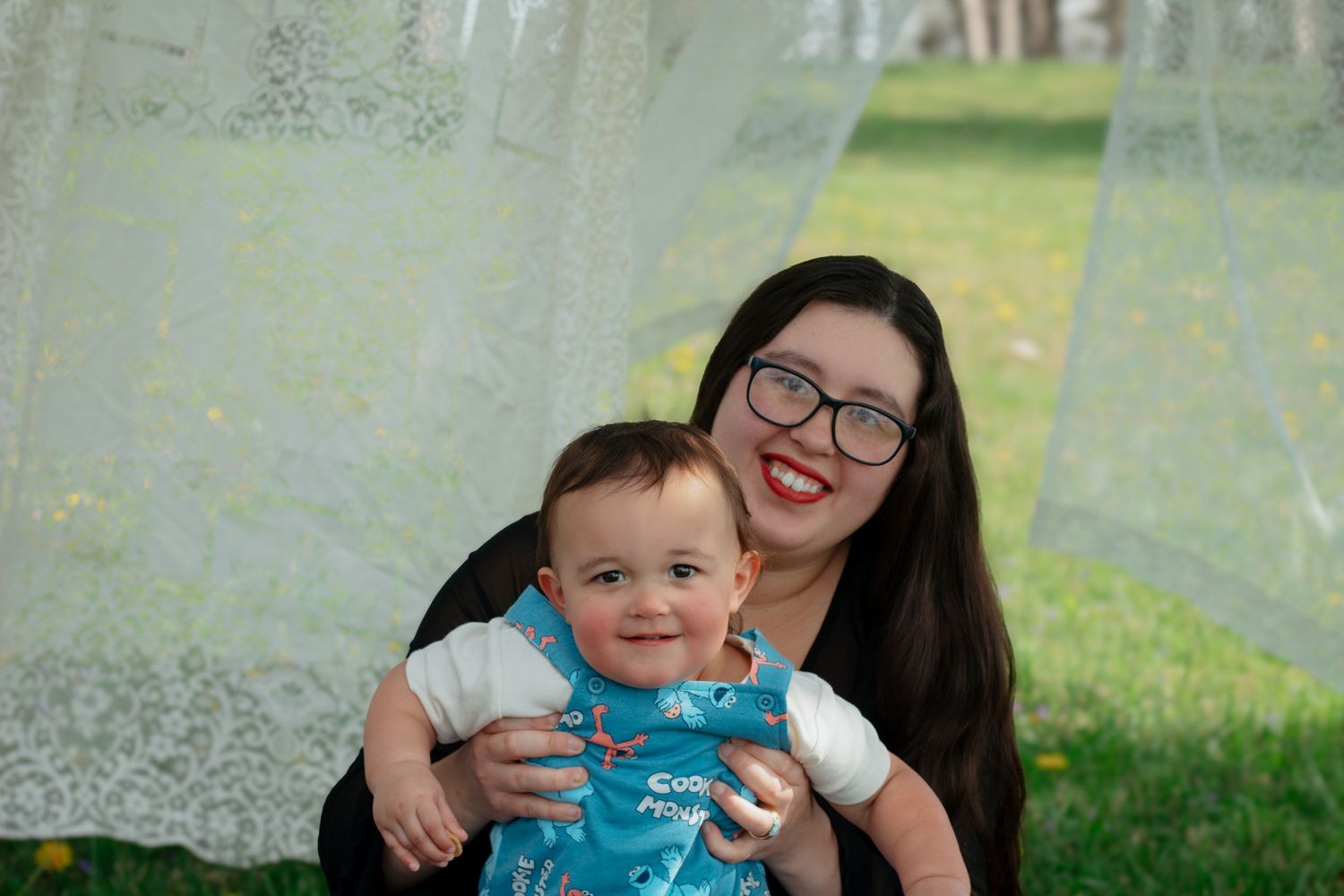 A smiling young woman with glasses holds a happy baby boy outdoors near lace curtains and green grass.